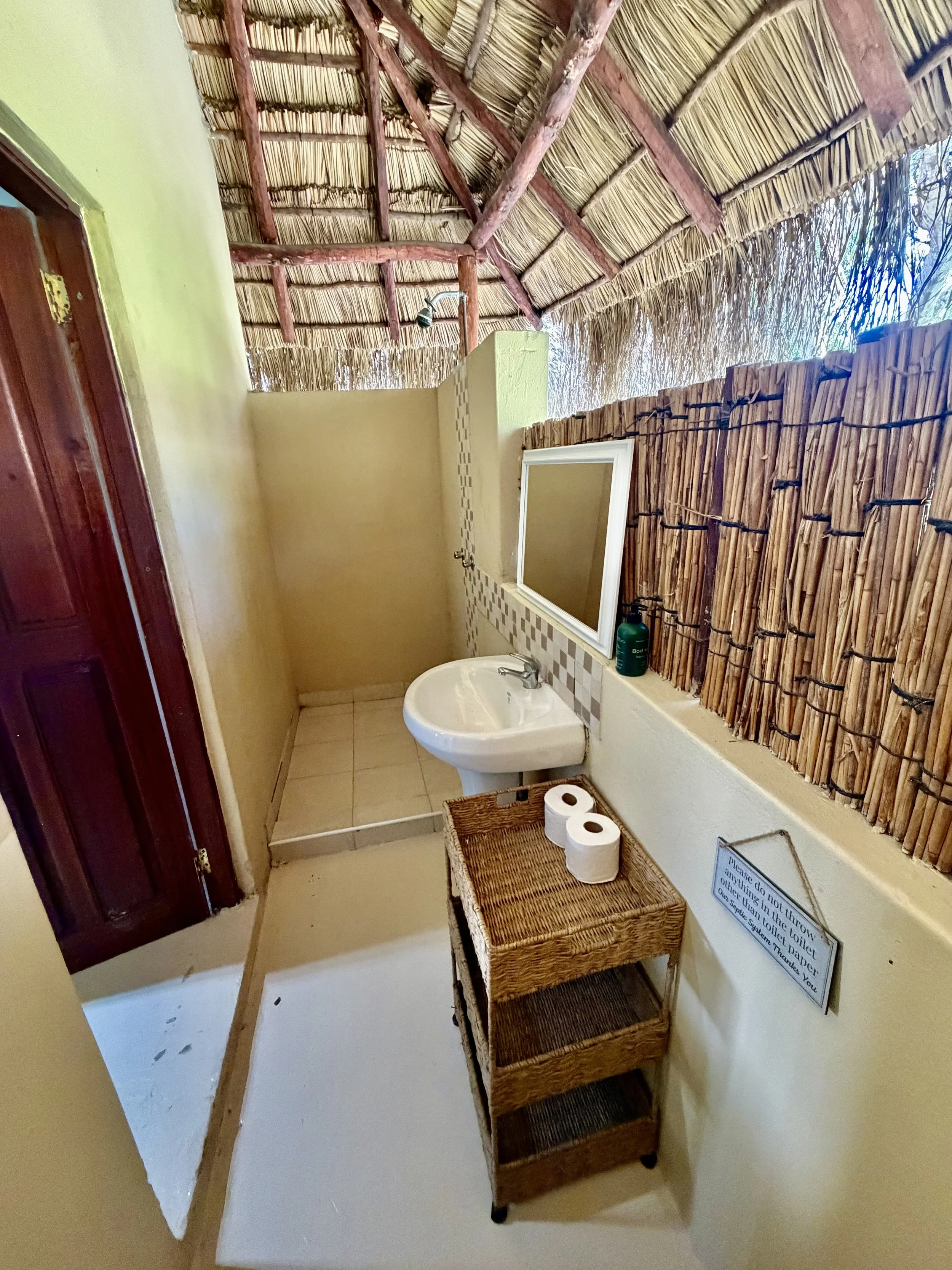 Bathroom with a tiki-style bamboo and thatch roof, beige painted walls, a small mirror above a sink, and a wooden shelf holding two rolls of toilet paper. There is a small sign near the shelf.