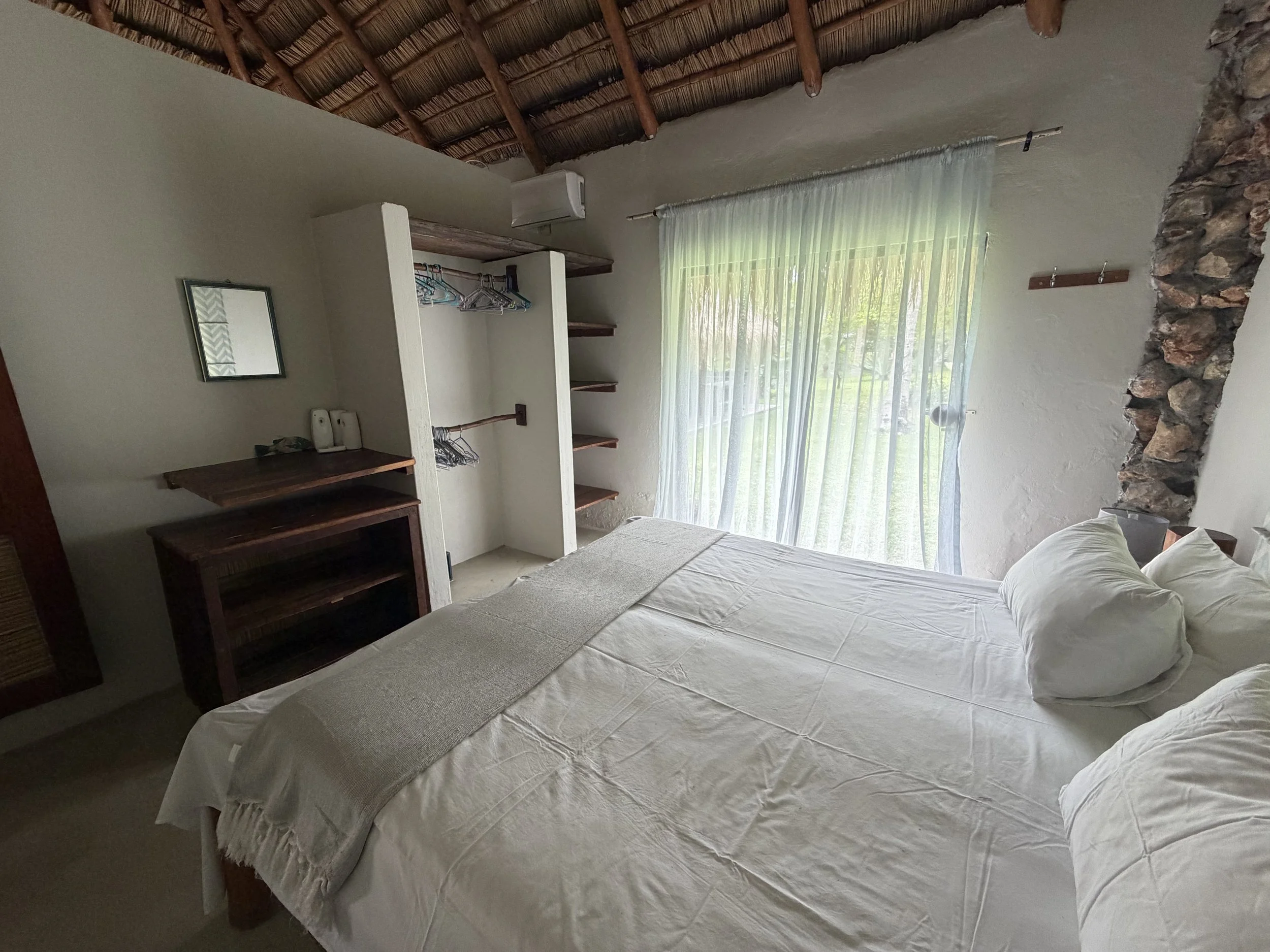 A cozy bedroom with a white bed, a stone accent wall, sheer curtains over large windows, a rustic wooden shelf, and a thatched roof ceiling.