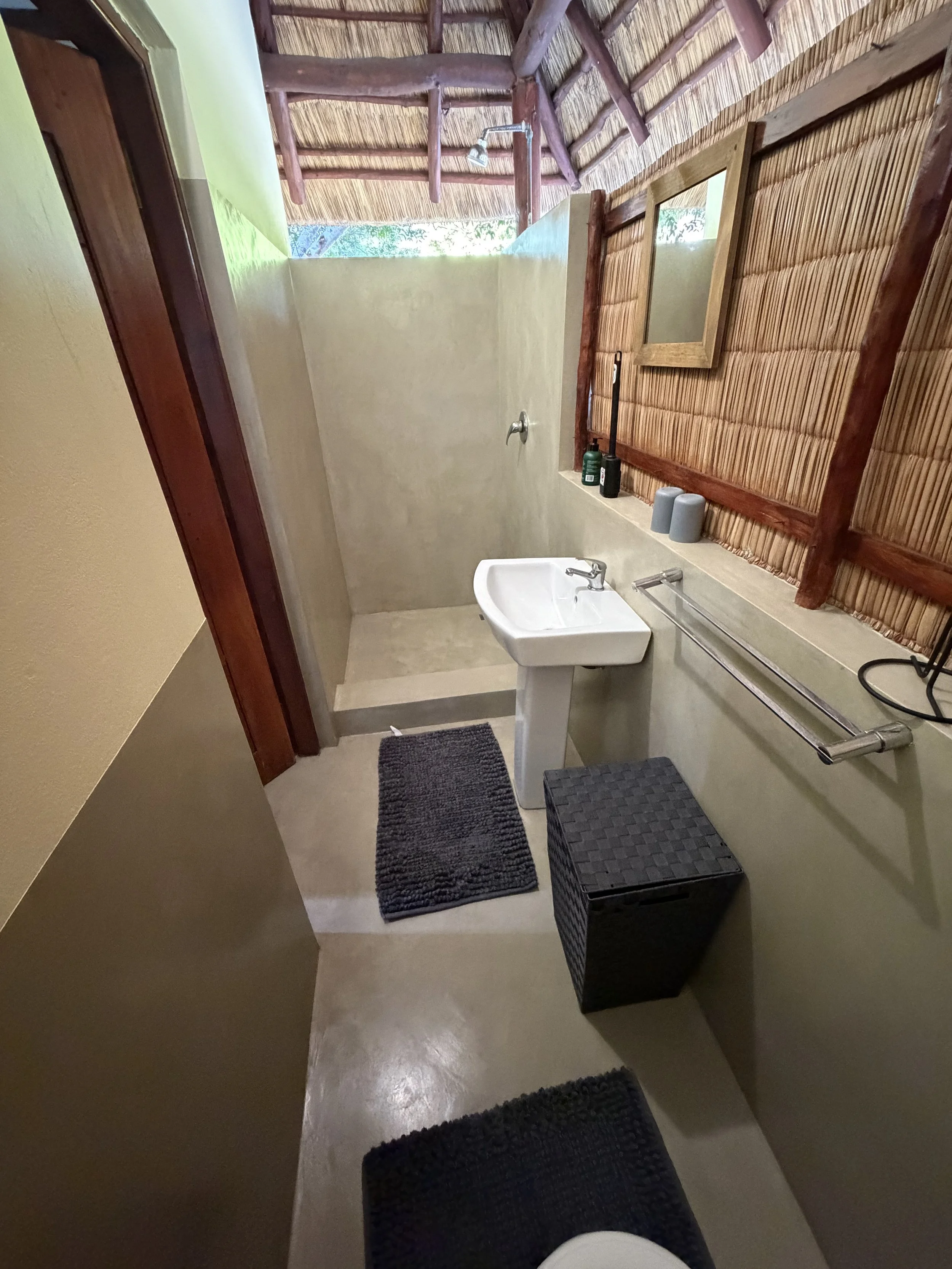 A rustic bathroom with a small sink, two gray cups, a black bottle of soap, a black woven basket, brown wooden trim, a mirror, and an outdoor shower with a bamboo roof and windows.