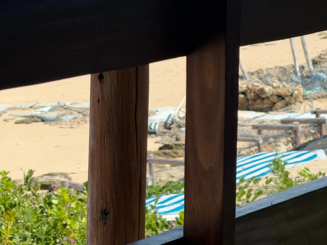 View through a wooden railing showing a sandy beach with blue and white striped umbrellas and some greenery.
