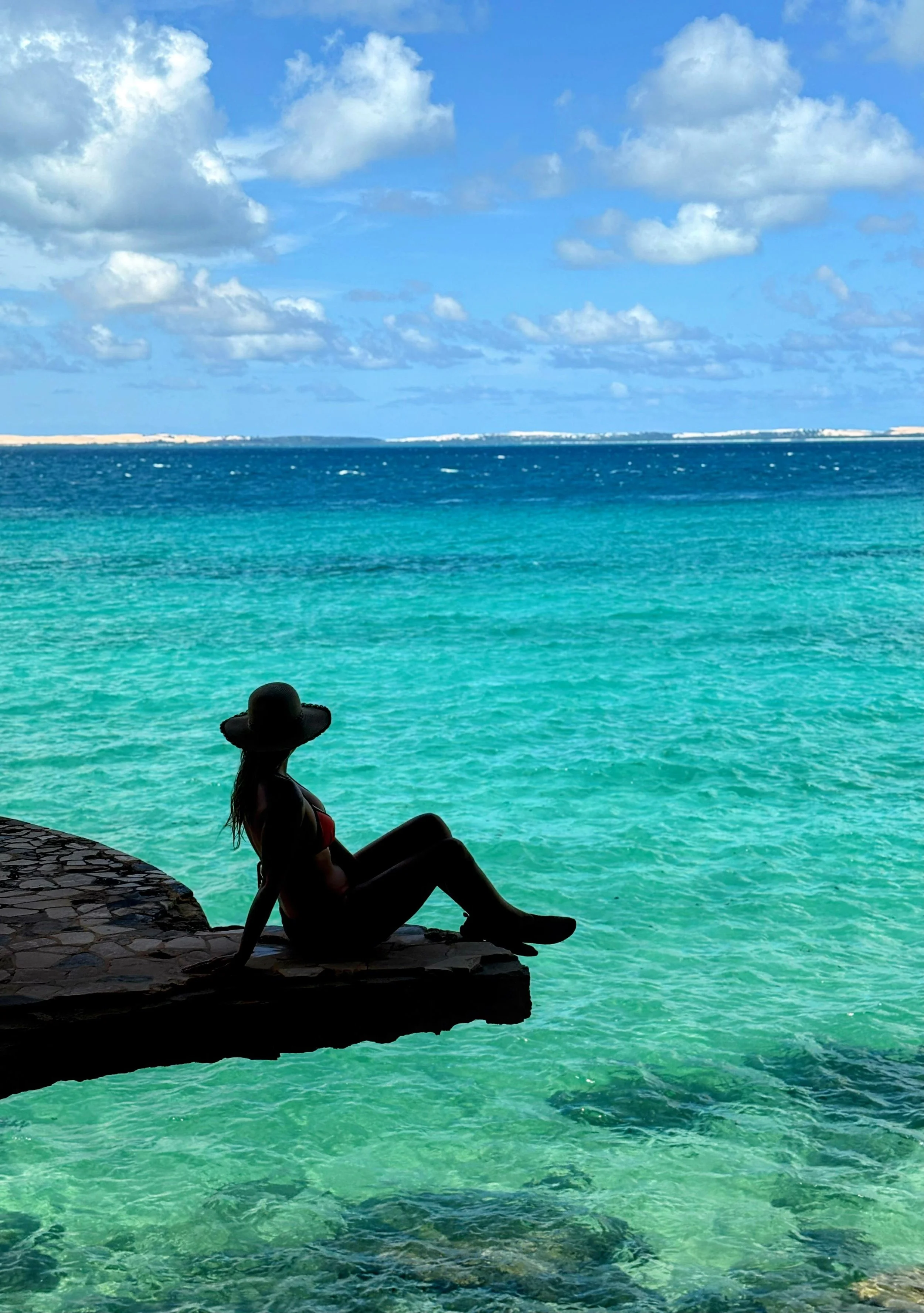 Person sitting on a rock ledge by turquoise water, wearing a wide-brimmed hat, with a scenic ocean and blue sky with clouds in the background.