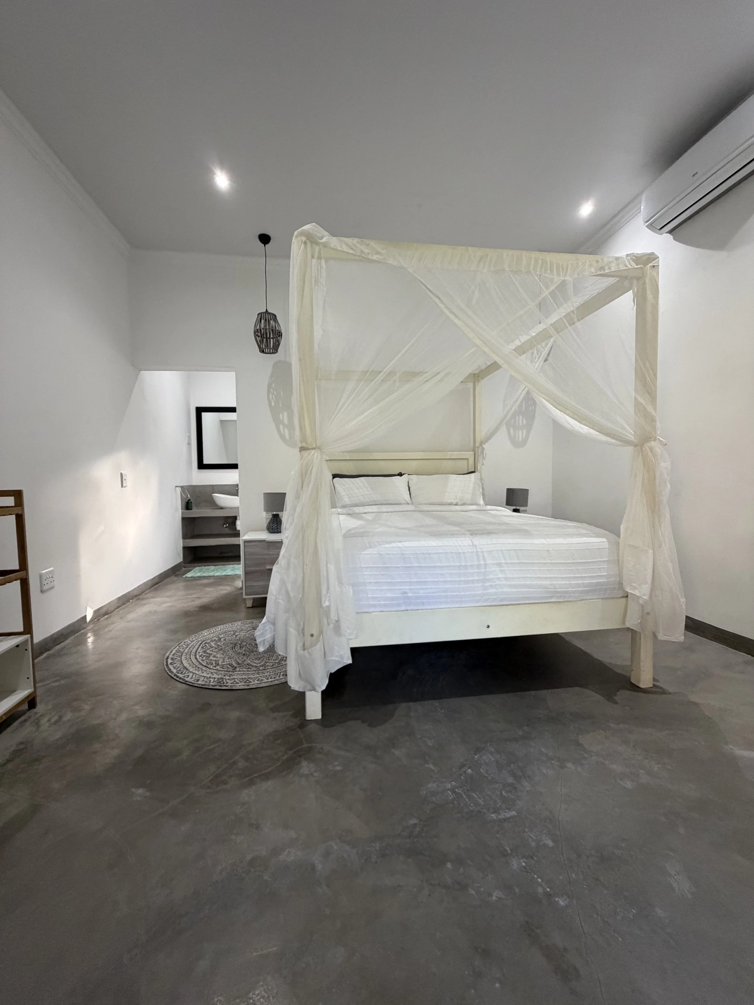 A white four-poster bed with sheer curtains in a modern bedroom. The room has white walls, a grey cement floor, a small bedside table with a lamp, a round patterned rug, a wall-mounted air conditioner, a hanging pendant light, and a bathroom sink visible in the background.