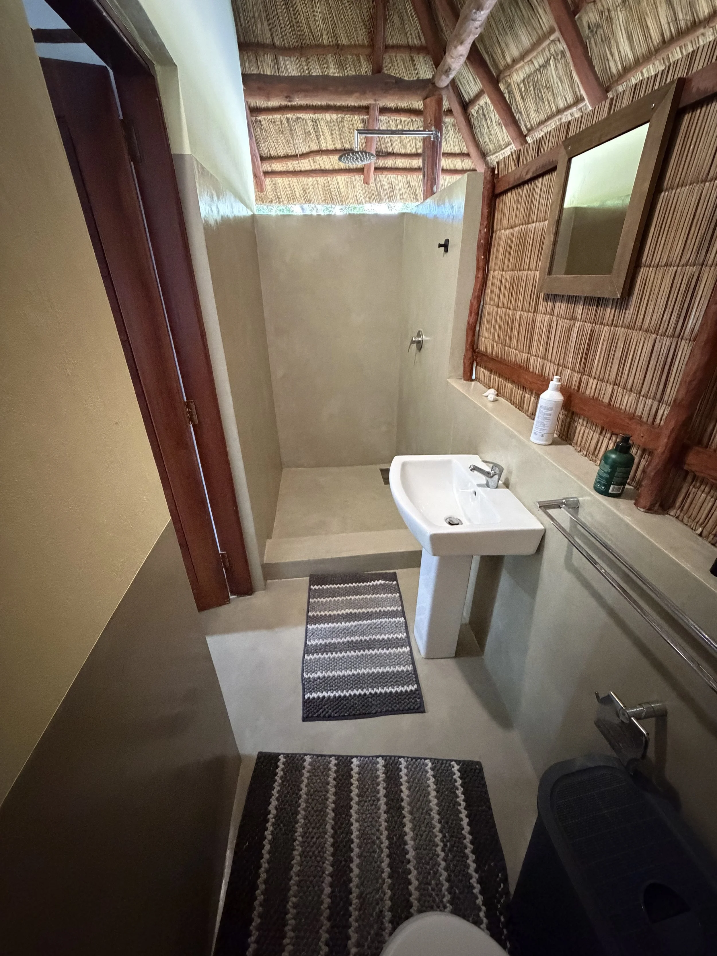 Bathroom with rustic decor, including bamboo wall coverings, a small sink, a mirror, a striped rug, and an outdoor shower with a thatched roof.