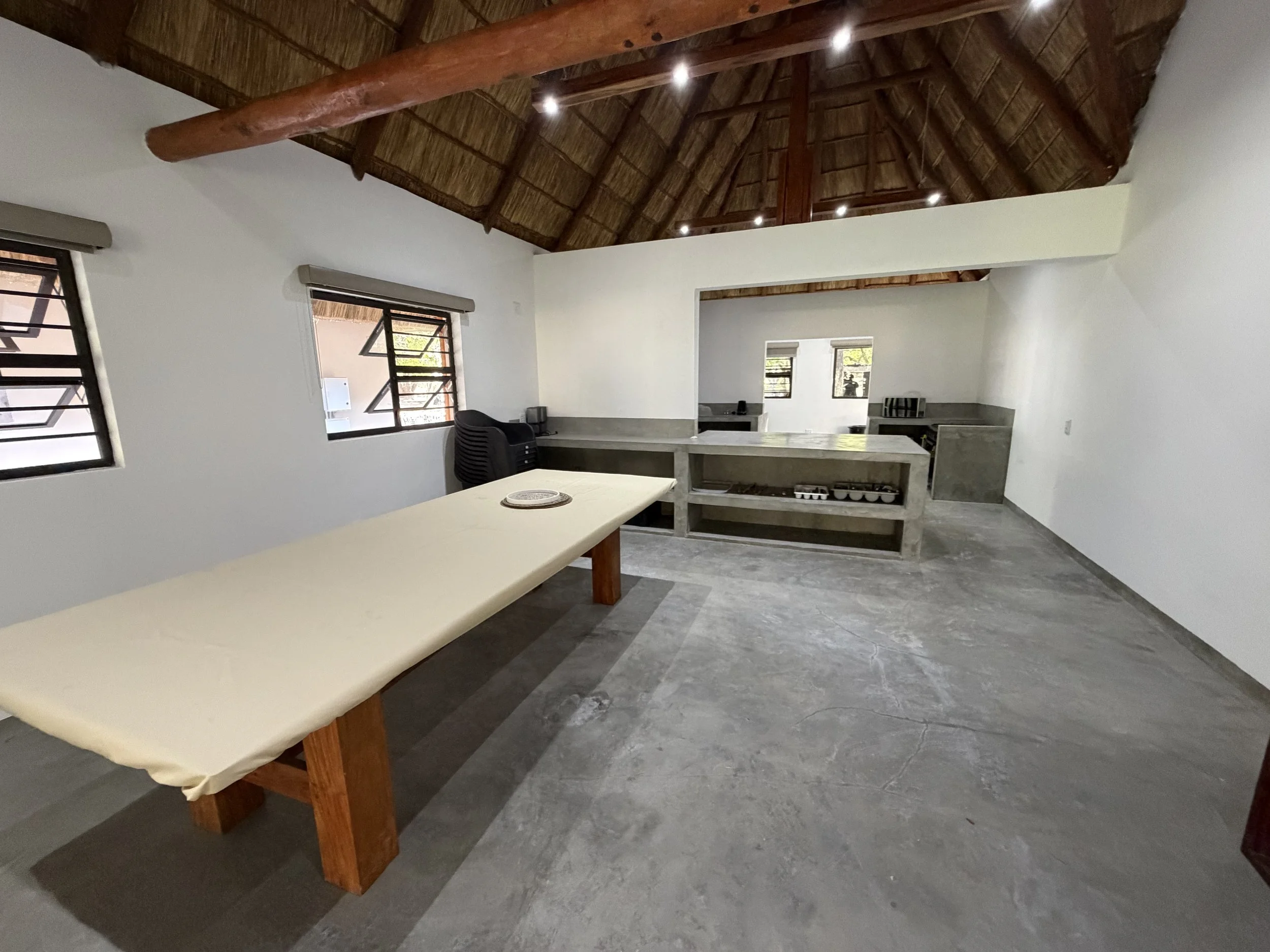 Interior of a room with a high thatched ceiling, white walls, and stone flooring. There is a rectangular table with a white top and wooden legs, a black chair, and a concrete kitchen counter with a stove. Three windows with metal bars and open louvered shutters are visible.