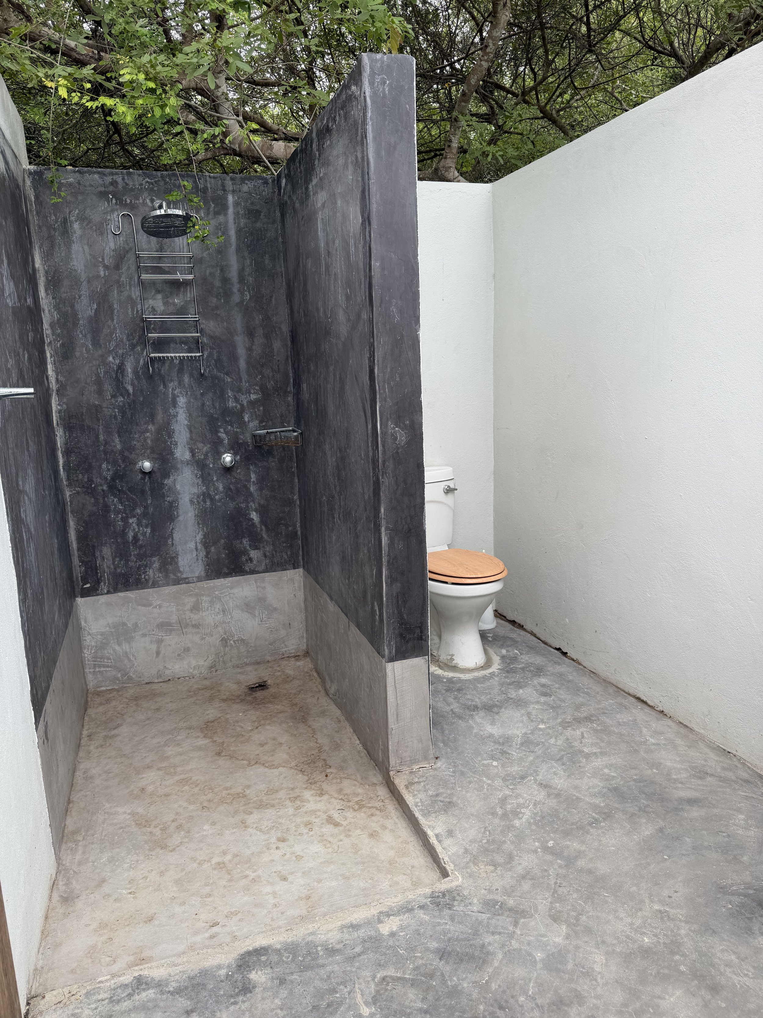 An outdoor restroom with a black shower on the left and a white toilet with a wooden seat on the right, separated by a low wall, with trees and greenery visible overhead.