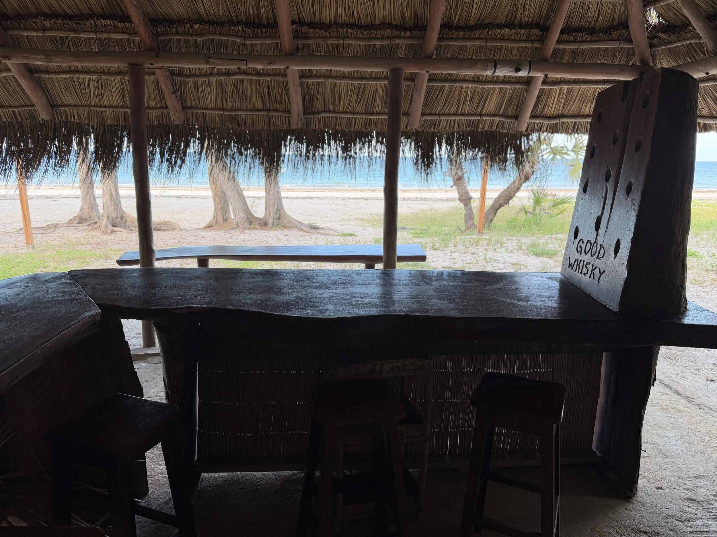 View from inside a thatched beach hut overlooking trees, sand, and ocean with a sign that says 'Good Whisky' on a wooden plaque.