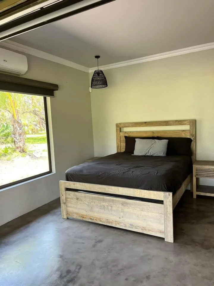A bedroom with a bed, black bedding, wooden headboard and side table, a window with view of trees, and a ceiling light fixture.