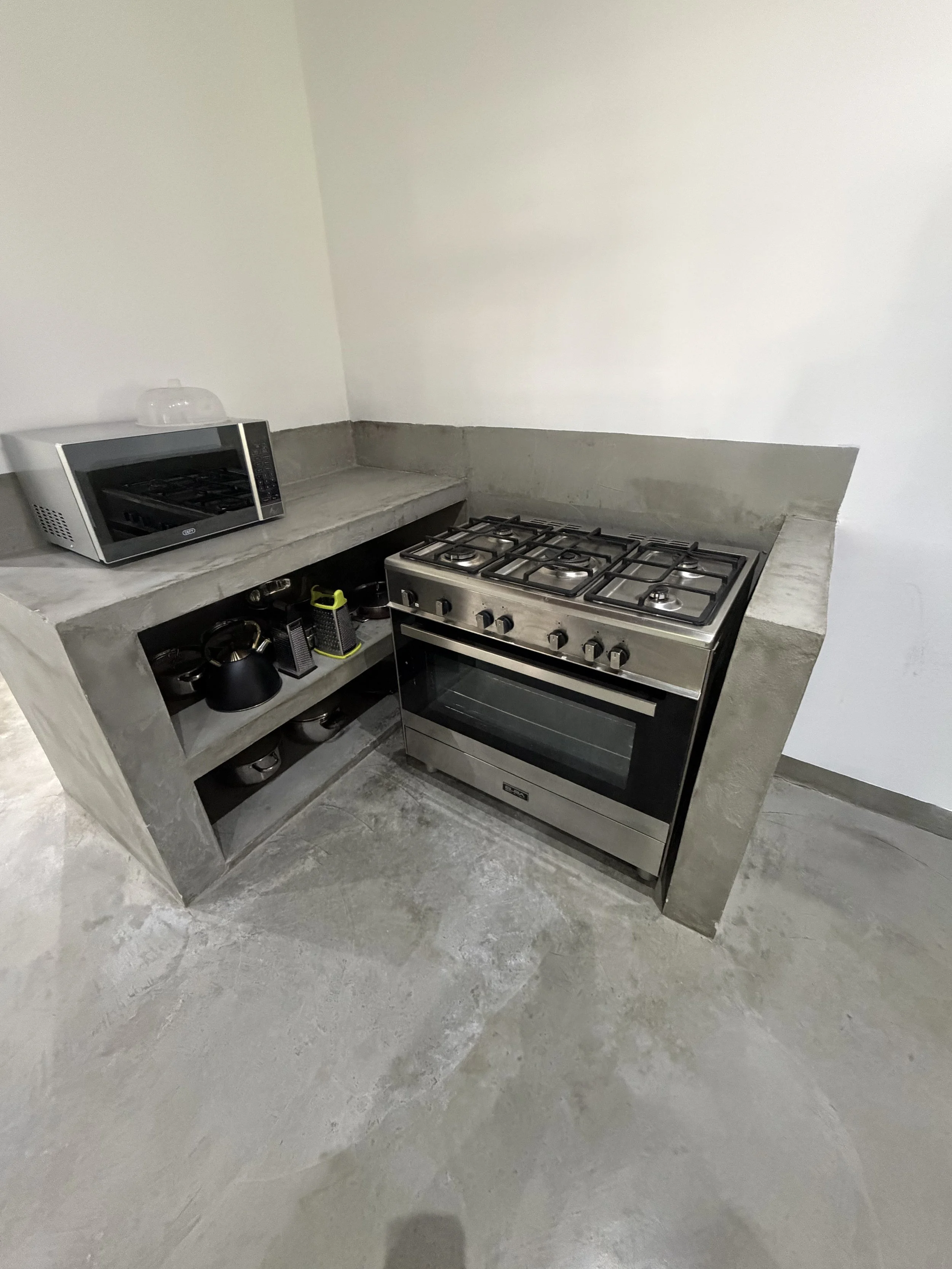 Unfinished kitchen with concrete countertops, a stove, a microwave, and shelving with kitchenware on a concrete floor.