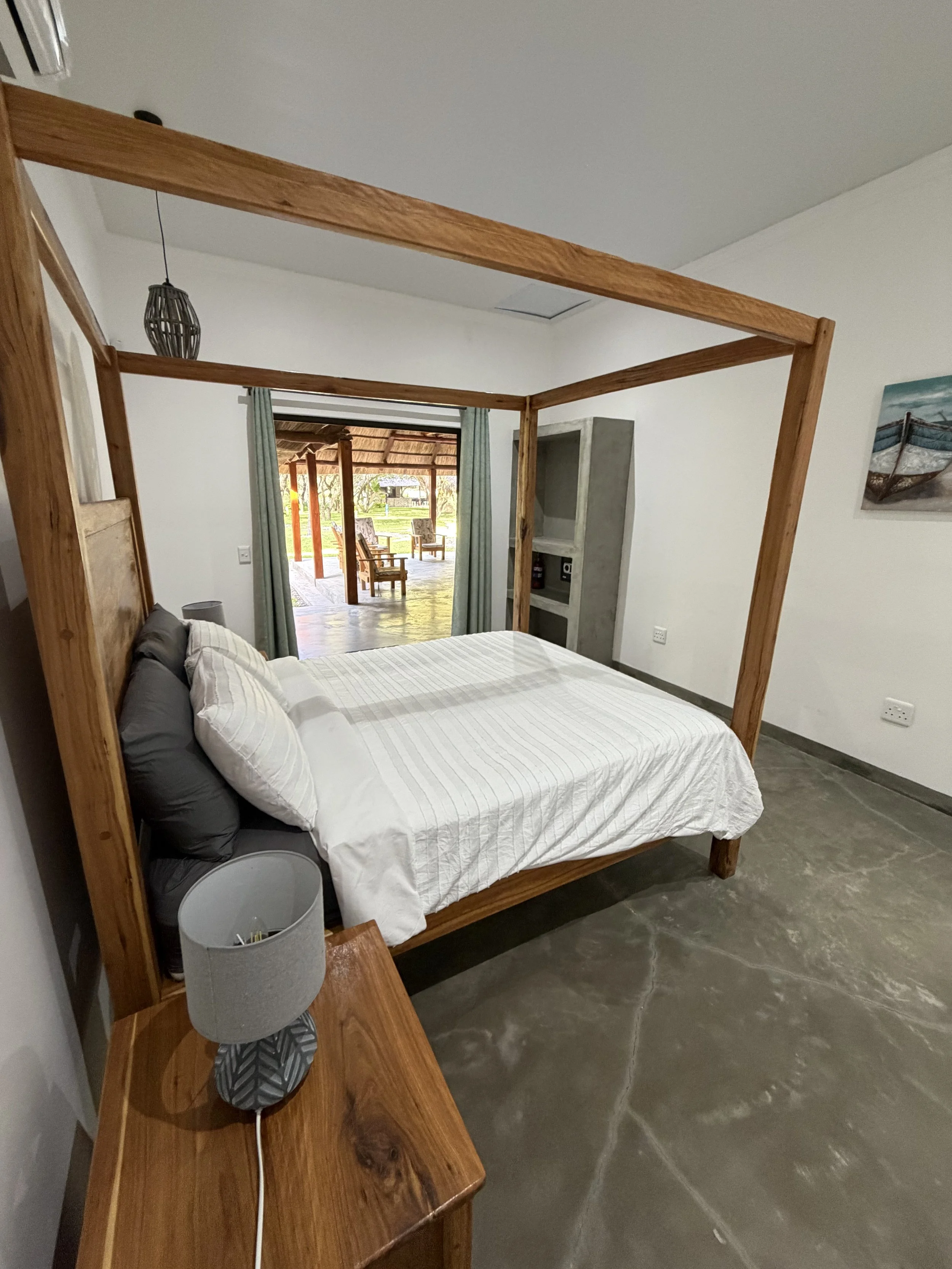 A bedroom with a wooden four-poster bed, white bedding, and pillows. A bedside table with a lamp and an electrical cord. An outdoor patio with chairs and a view of greenery through sliding glass doors.
