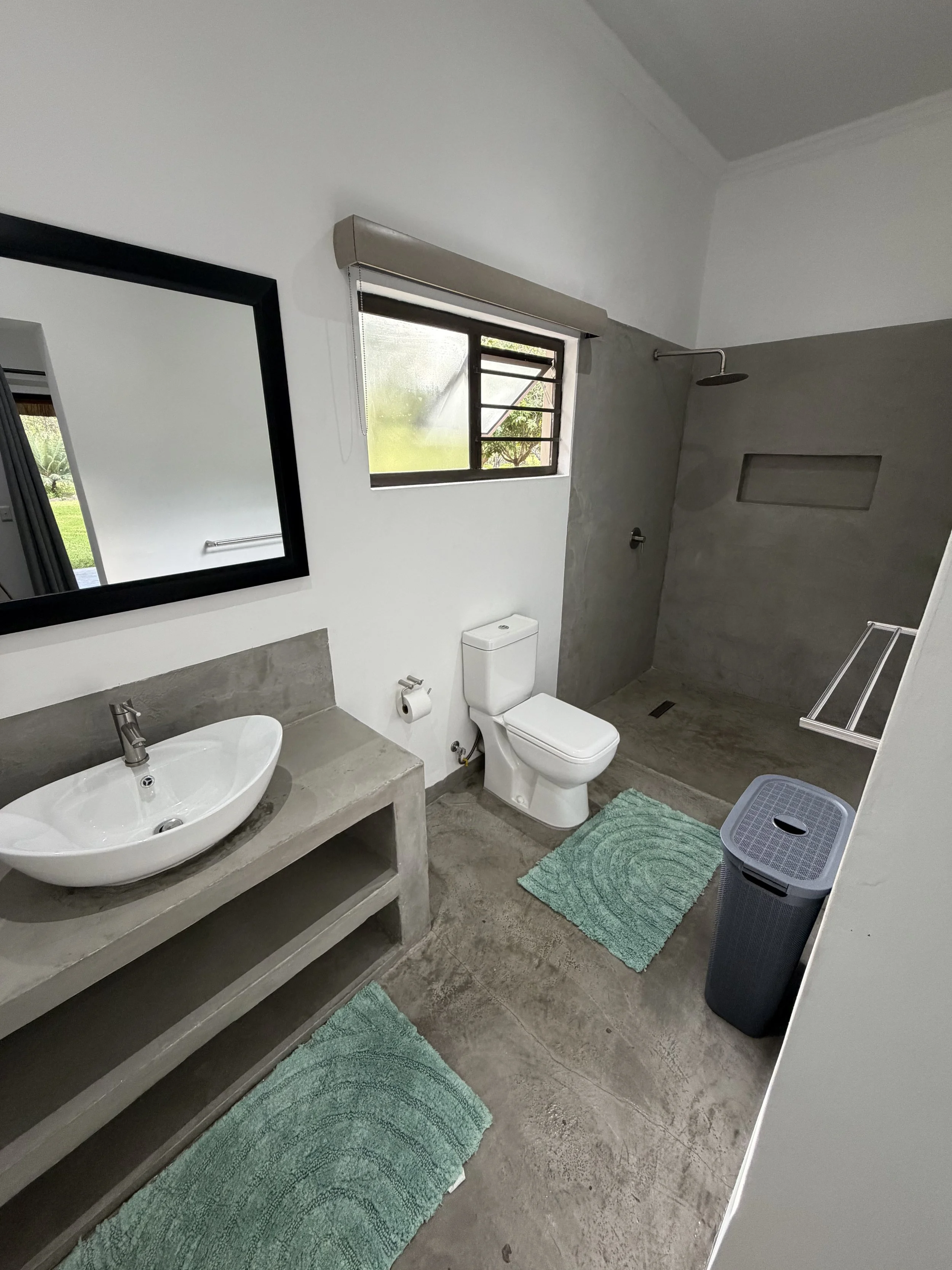 Modern bathroom with a concrete sink, a mirror, a toilet, a shower area with a rain showerhead, a small window, a power outlet, a laundry basket, and green bath mats.