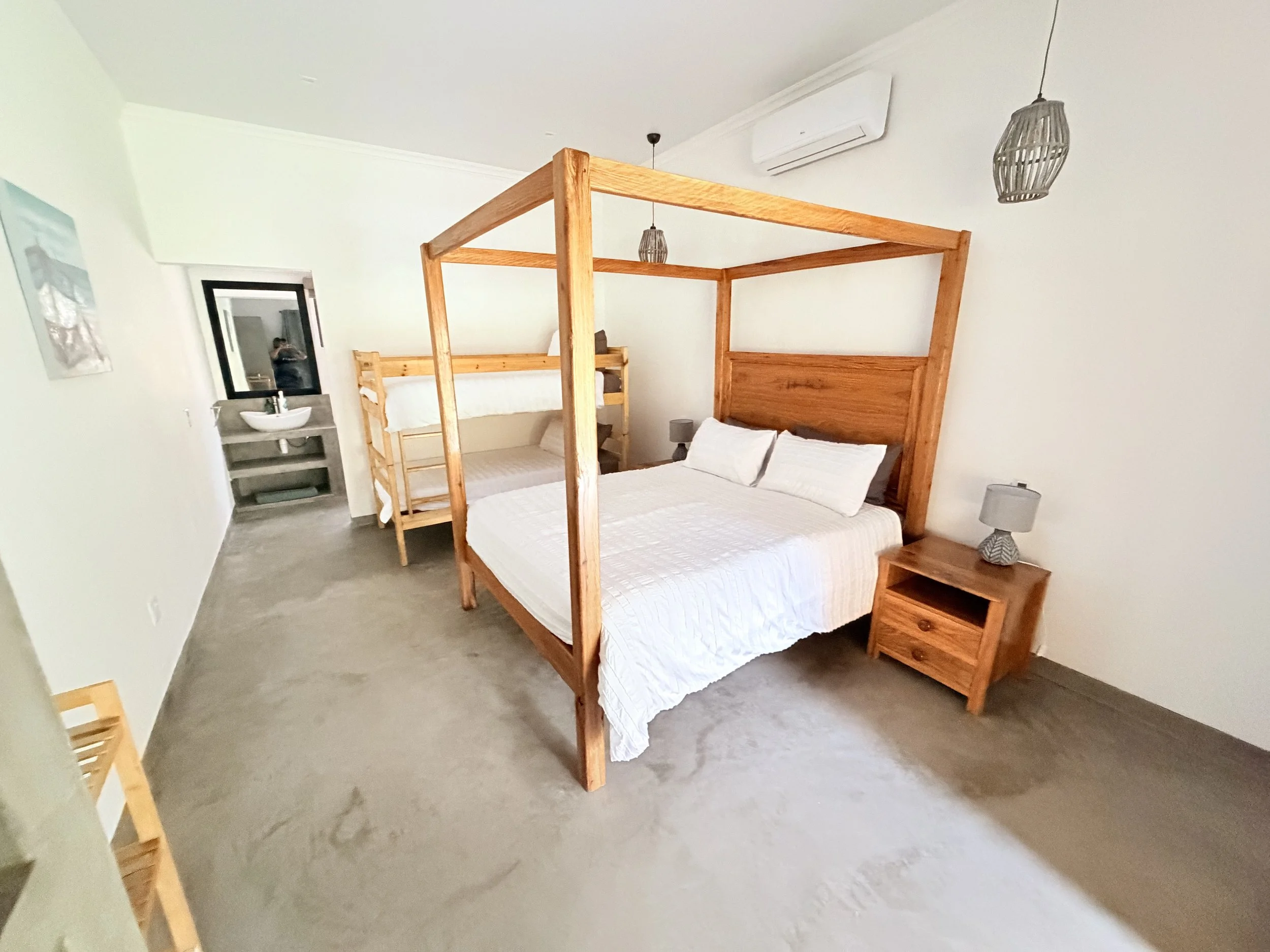 A bedroom with a wooden four-poster bed with white bedding, two bedside tables with lamps, a small sitting area, and a bathroom sink visible through an open door. The room has white walls, a gray carpet, and ceiling lights with wicker shades.
