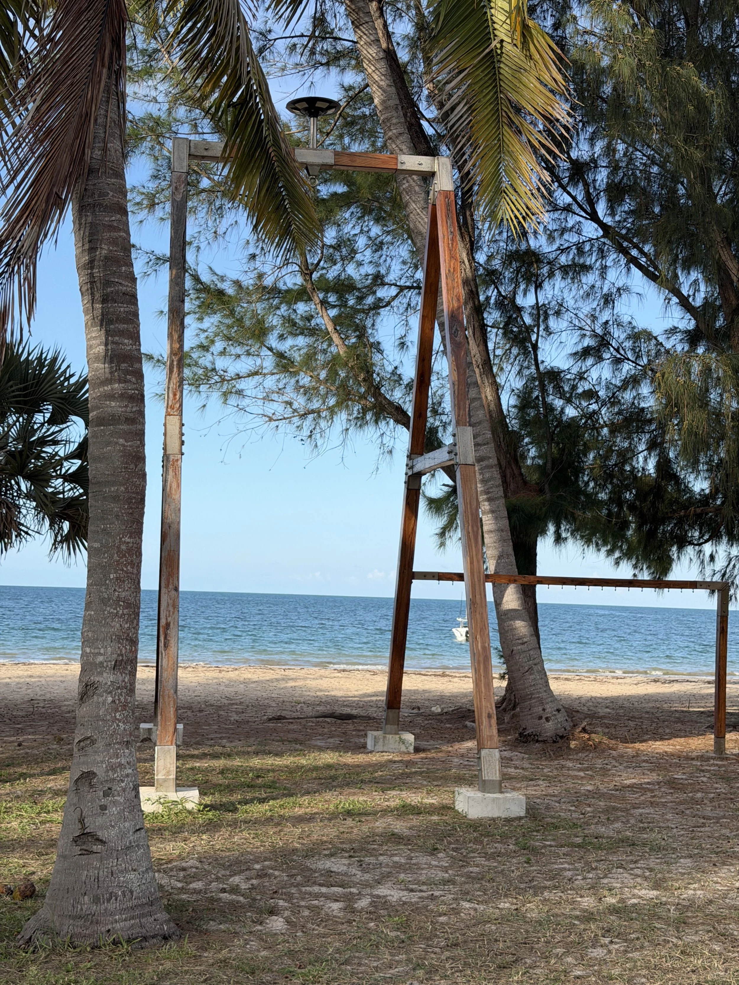 A swing set frame on a sandy beach with palm trees and the ocean in the background.