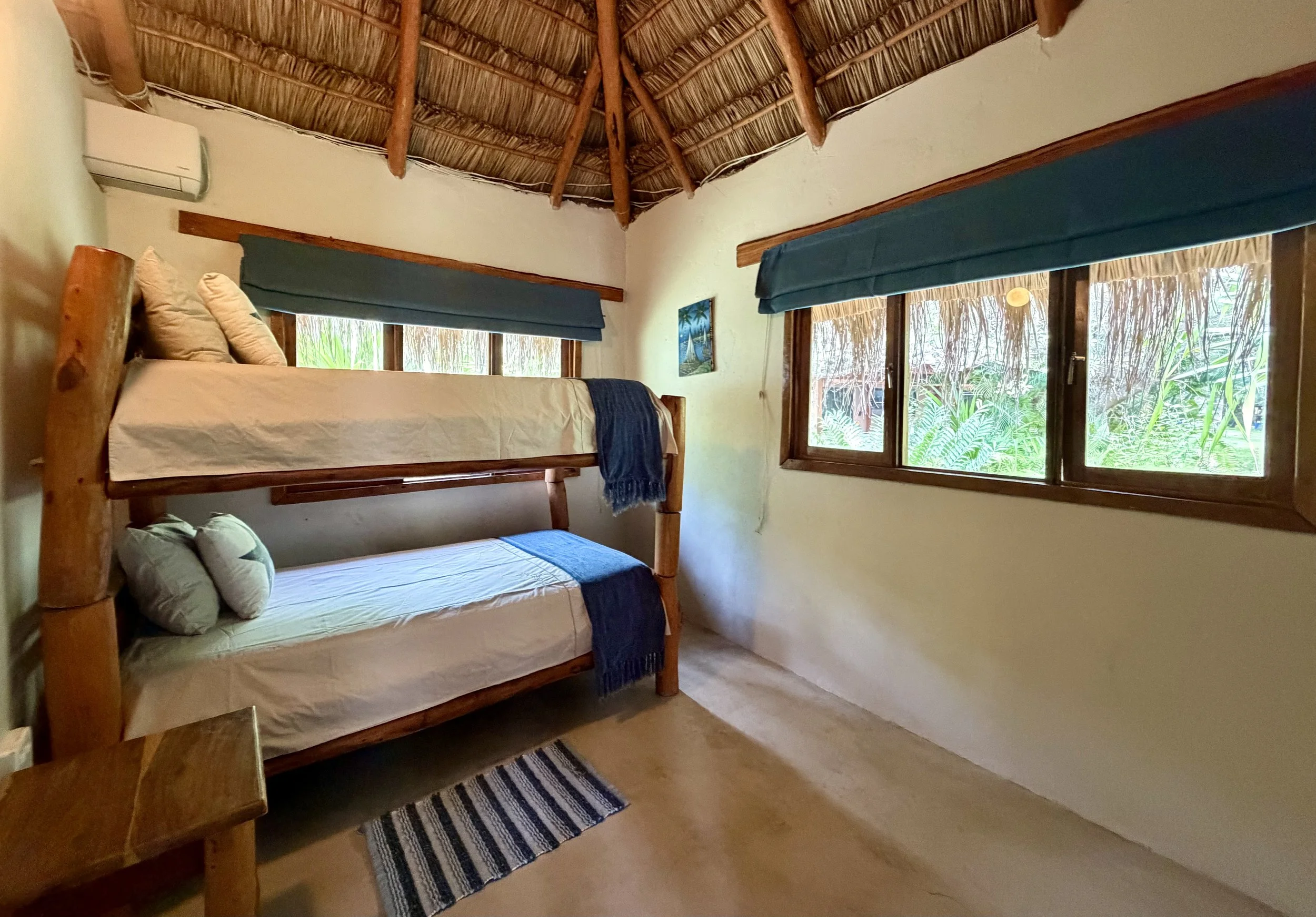 Bedroom with wooden bunk beds, blue window covers, tropical artwork, and lush greenery visible through the windows.