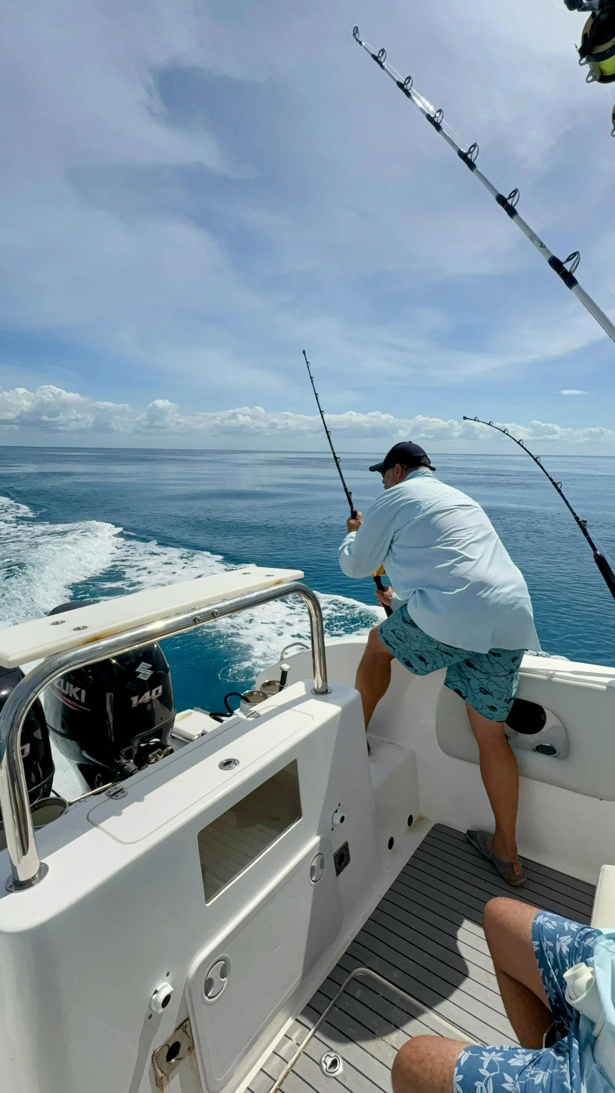 A man fishing off the side of a boat in the open ocean while another person sitting nearby watches, with a clear blue sky and some clouds in the background.