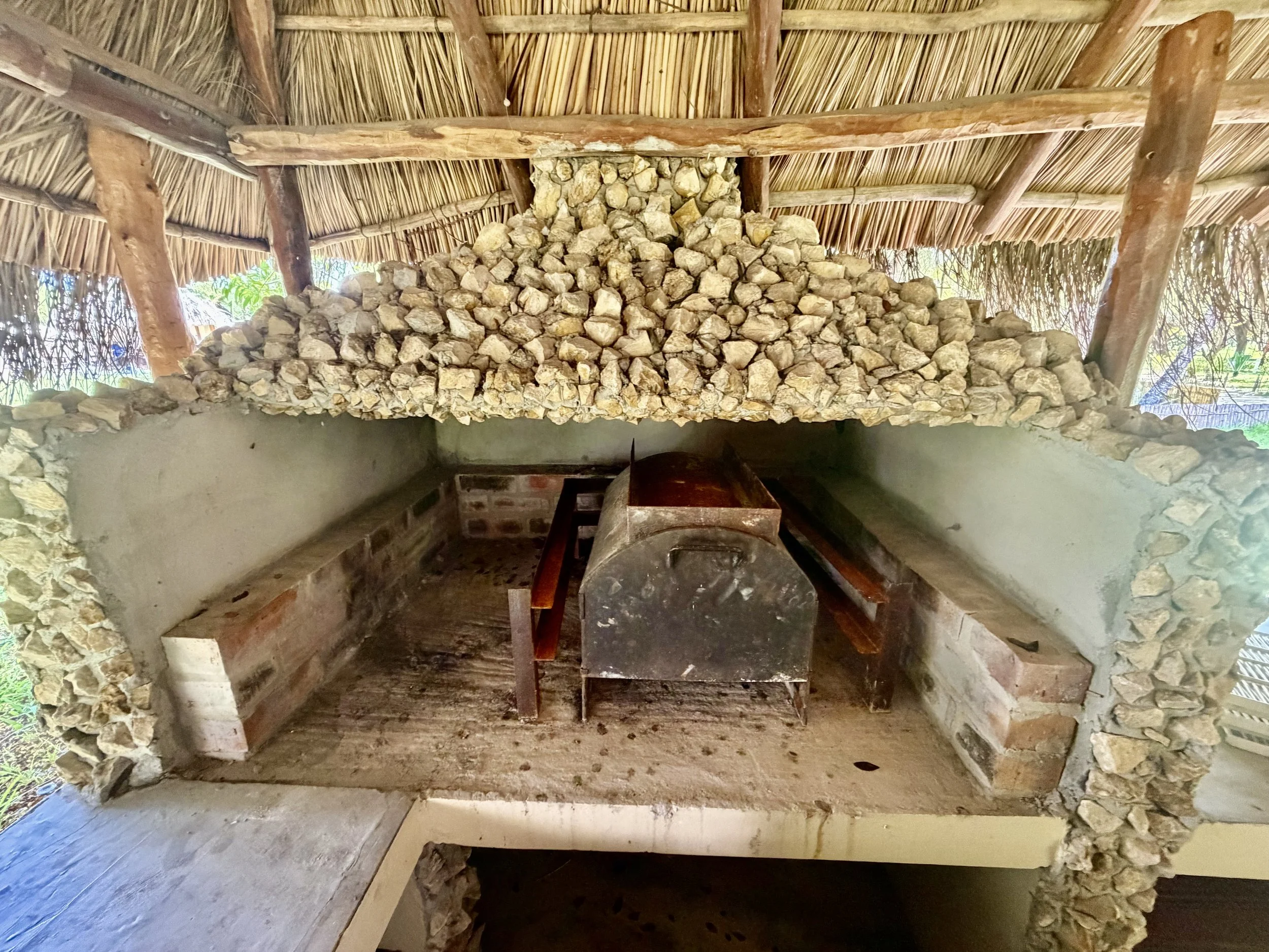 Thatched roof made of dry grass supported by wooden beams. Inside, a structure with stone and brick walls and a small wood-fired oven or stove.