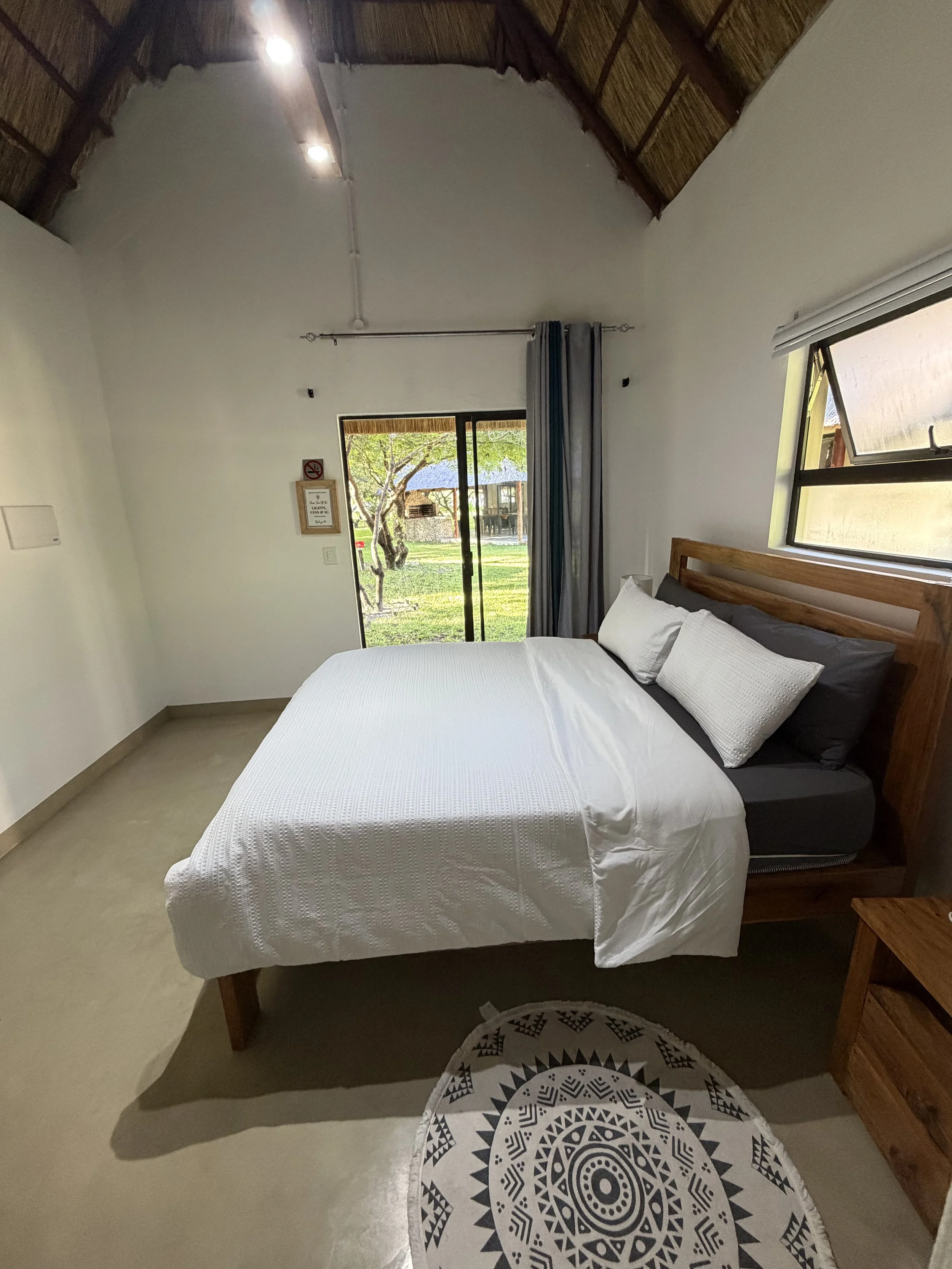 A bedroom with a bed covered with white bedding, a wooden headboard, and pillows. There is a round black and white patterned rug on the beige floor. Two windows, one with blinds and one with a curtain, let in natural light. Outside, there is a yard with a tree and a thatched-roof structure.