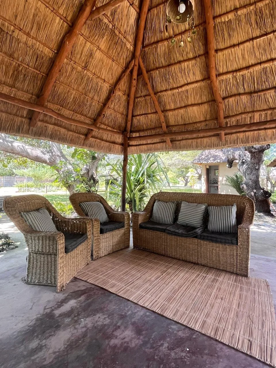 Wicker furniture set with three chairs and a sofa, each with striped pillows, placed on a woven rug under a thatched roof gazebo surrounded by trees and greenery.