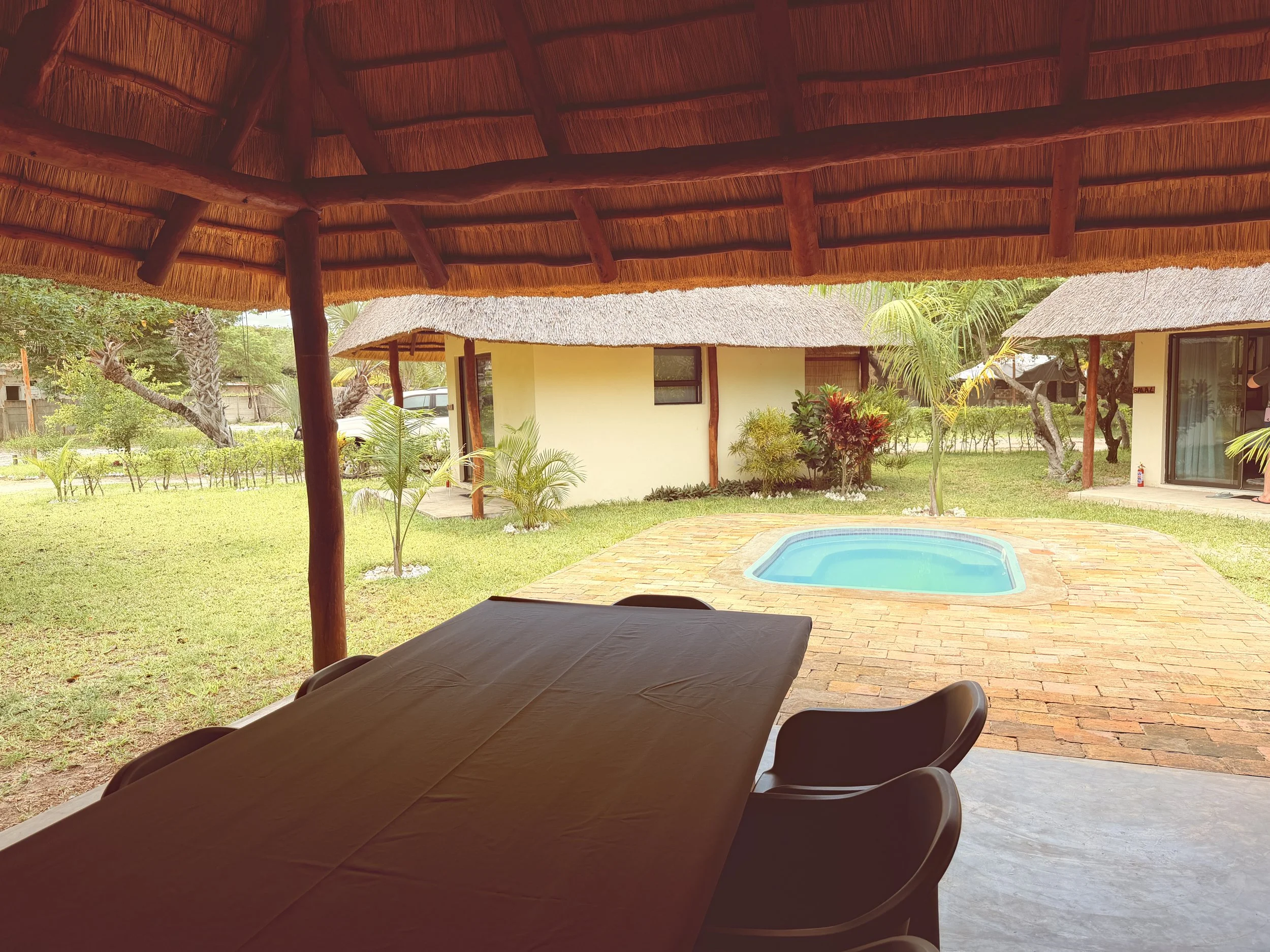 View from under a thatched roof patio looking out onto a yard with a small swimming pool, surrounded by yellow buildings, trees, and greenery.