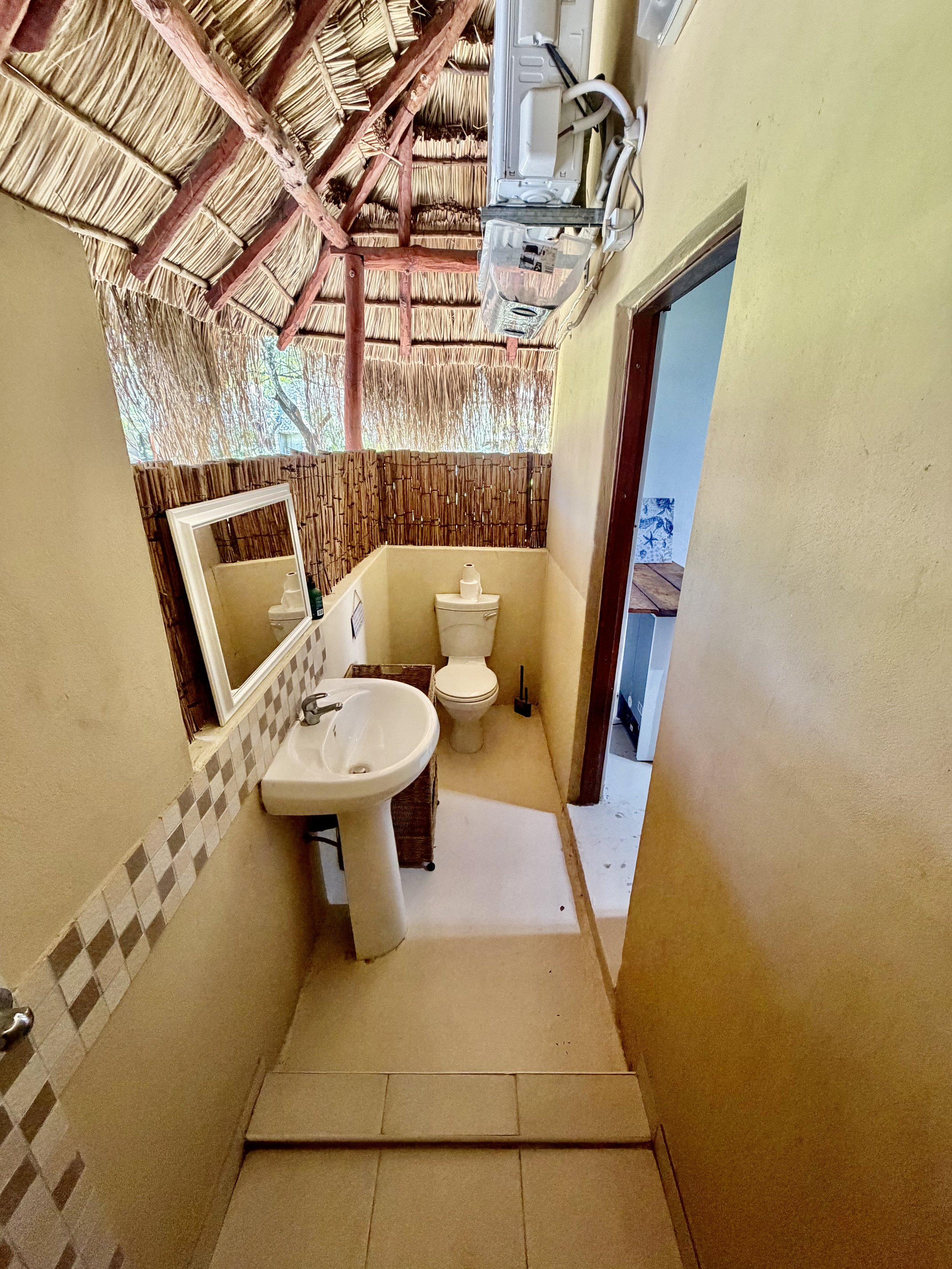 Small bathroom with a thatched roof ceiling, a toilet, a pedestal sink, a mirror, and a curtain of reeds dividing the space. An air conditioning unit is mounted on the wall.