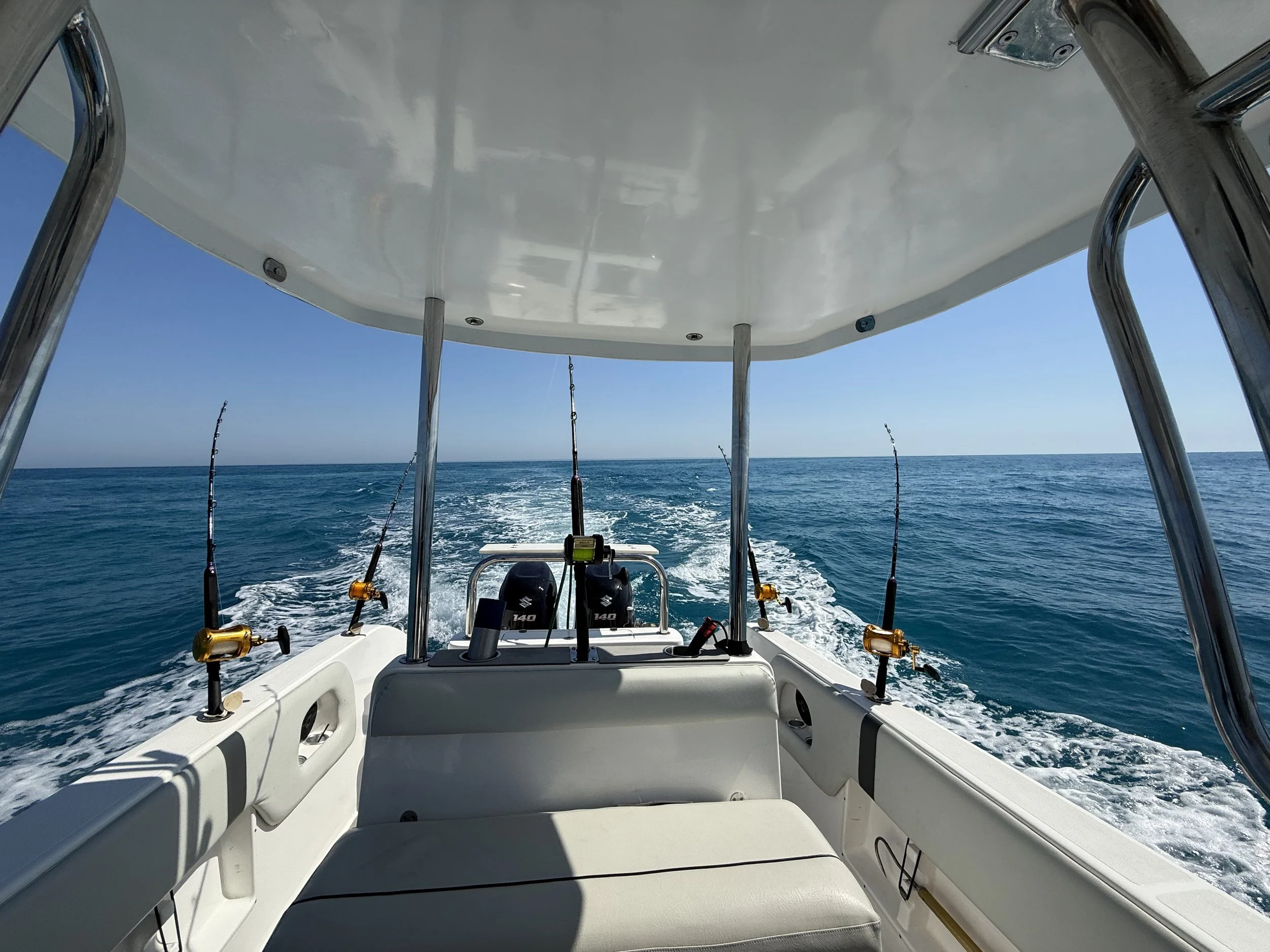 View from the back of a boat with fishing rods, heading out over calm ocean water under a clear blue sky.