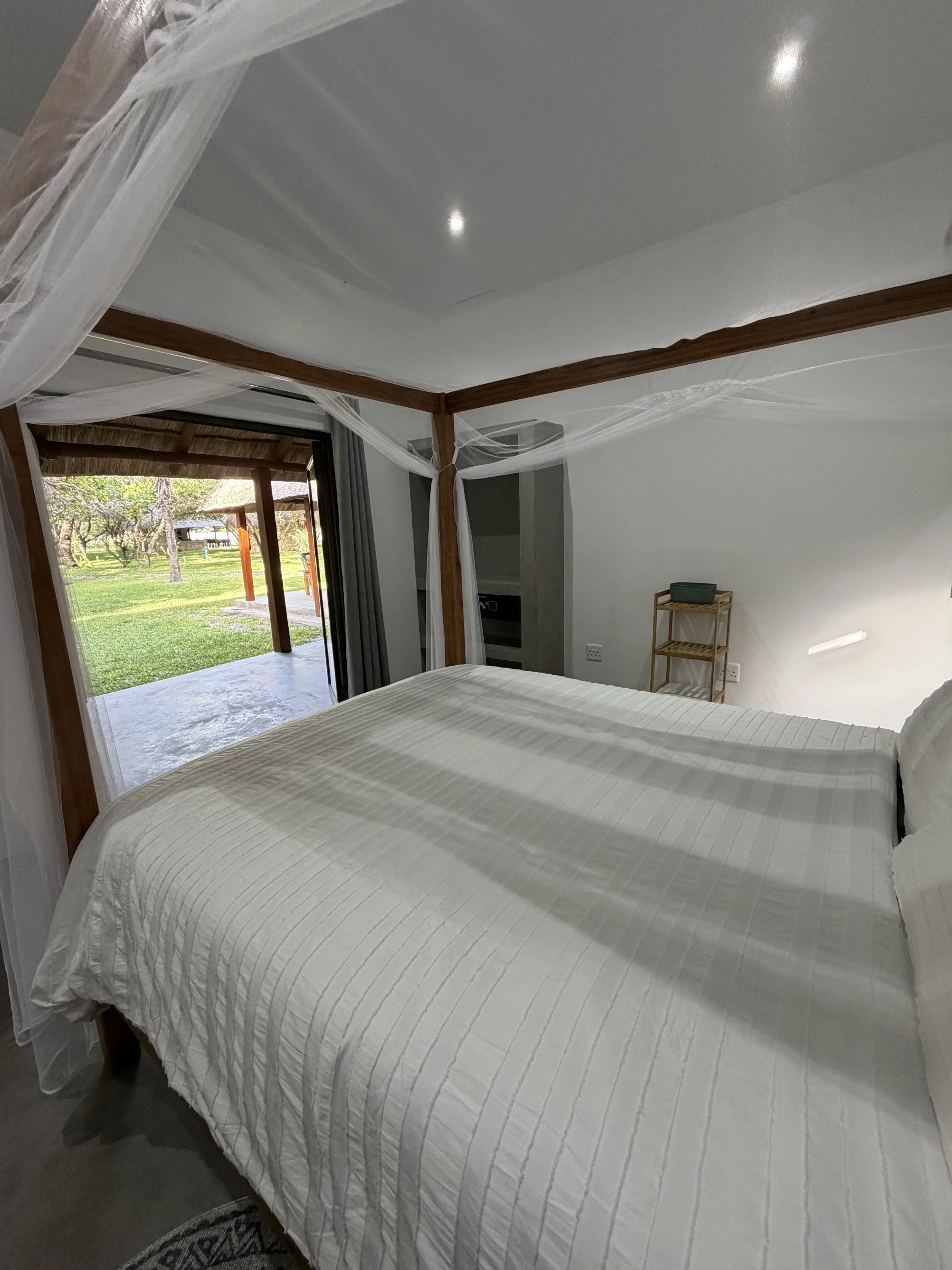 A bedroom with a bed covered in a white quilt, a wooden canopy frame with sheer curtains, a sliding door opening to an outdoor patio and grassy area, and a small wooden shelving unit with a green box on top.