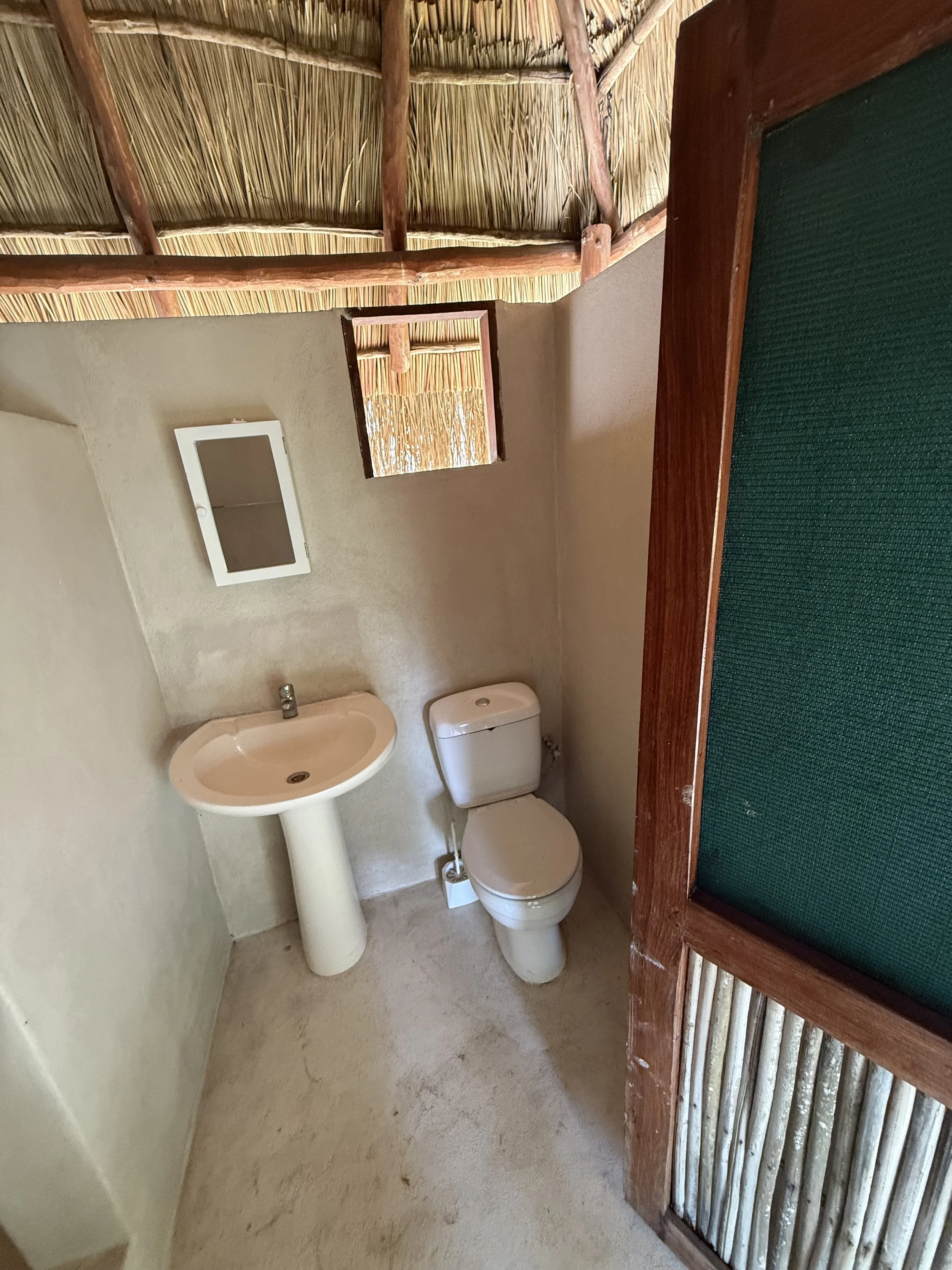 A small bathroom with a beige wall, a white sink, a white toilet, a small mirror, and a small window with woven bamboo shade, with part of a green-topped wooden structure in the foreground.