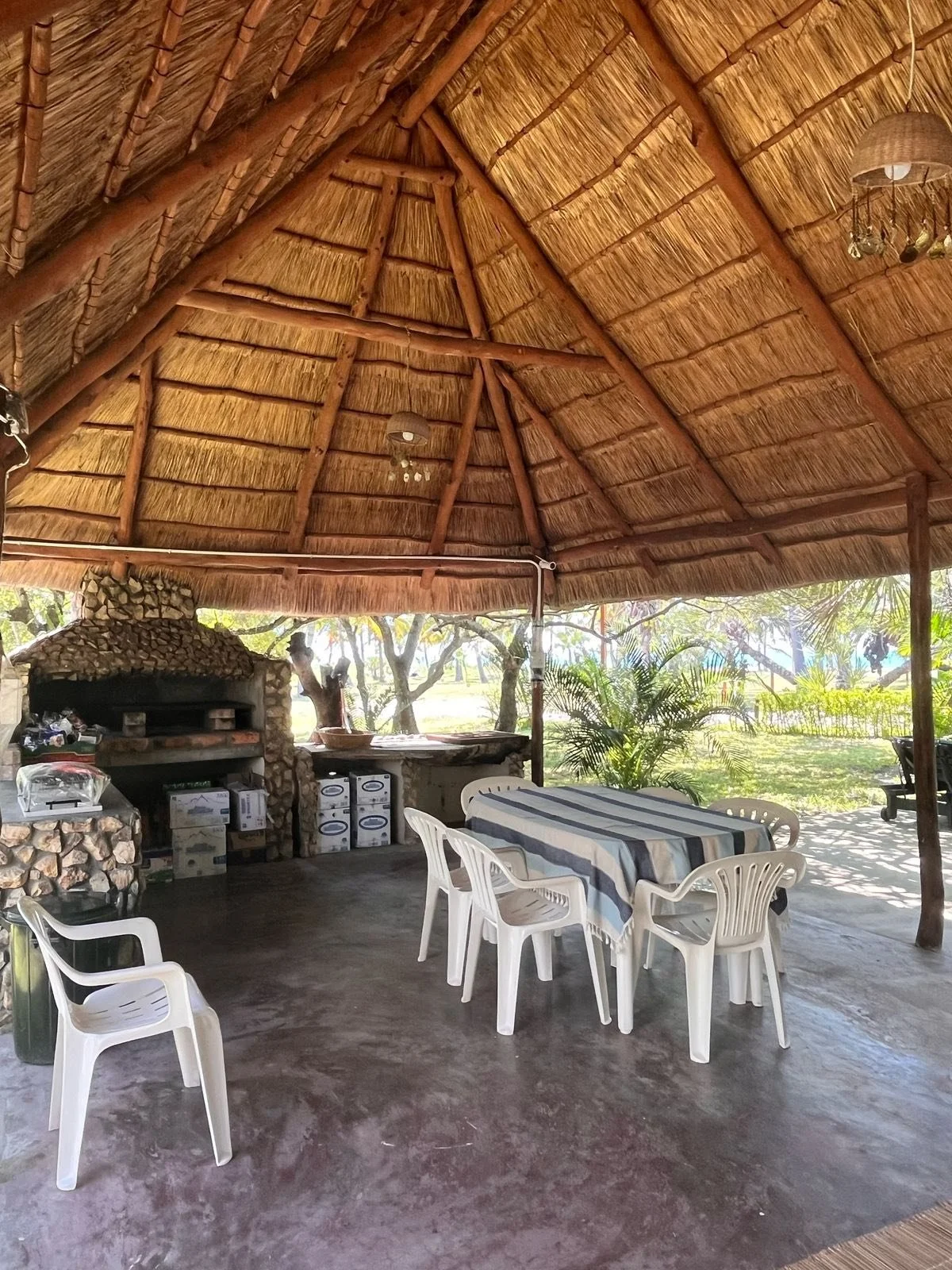 Outdoor dining area with a thatched roof, plastic chairs, a table with a striped tablecloth, and a stone pizza oven in the background surrounded by trees and greenery.