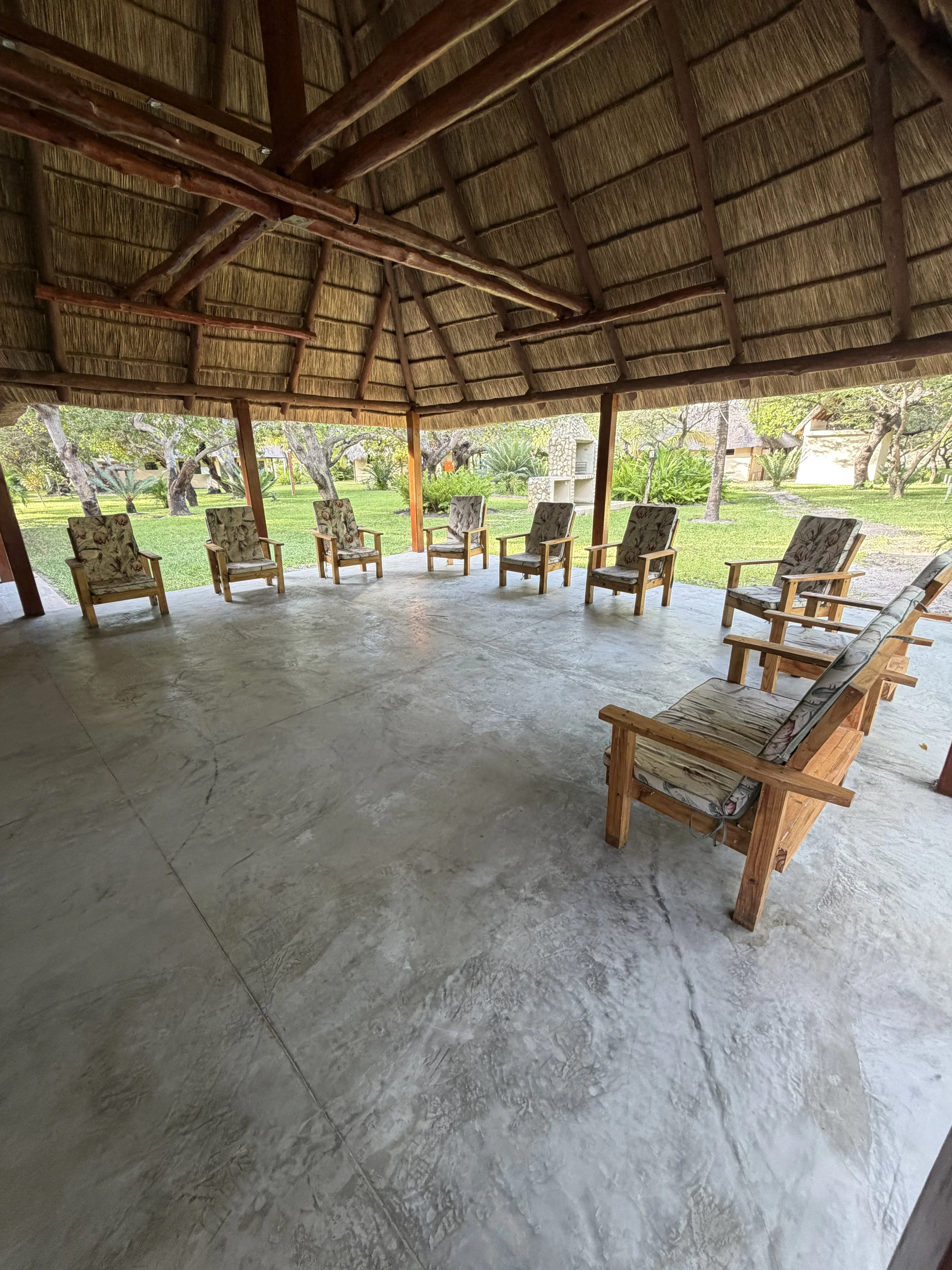 Open thatched-roof gazebo with eight wooden chairs with floral cushions around the perimeter, overlooking a lush garden with trees and bushes.