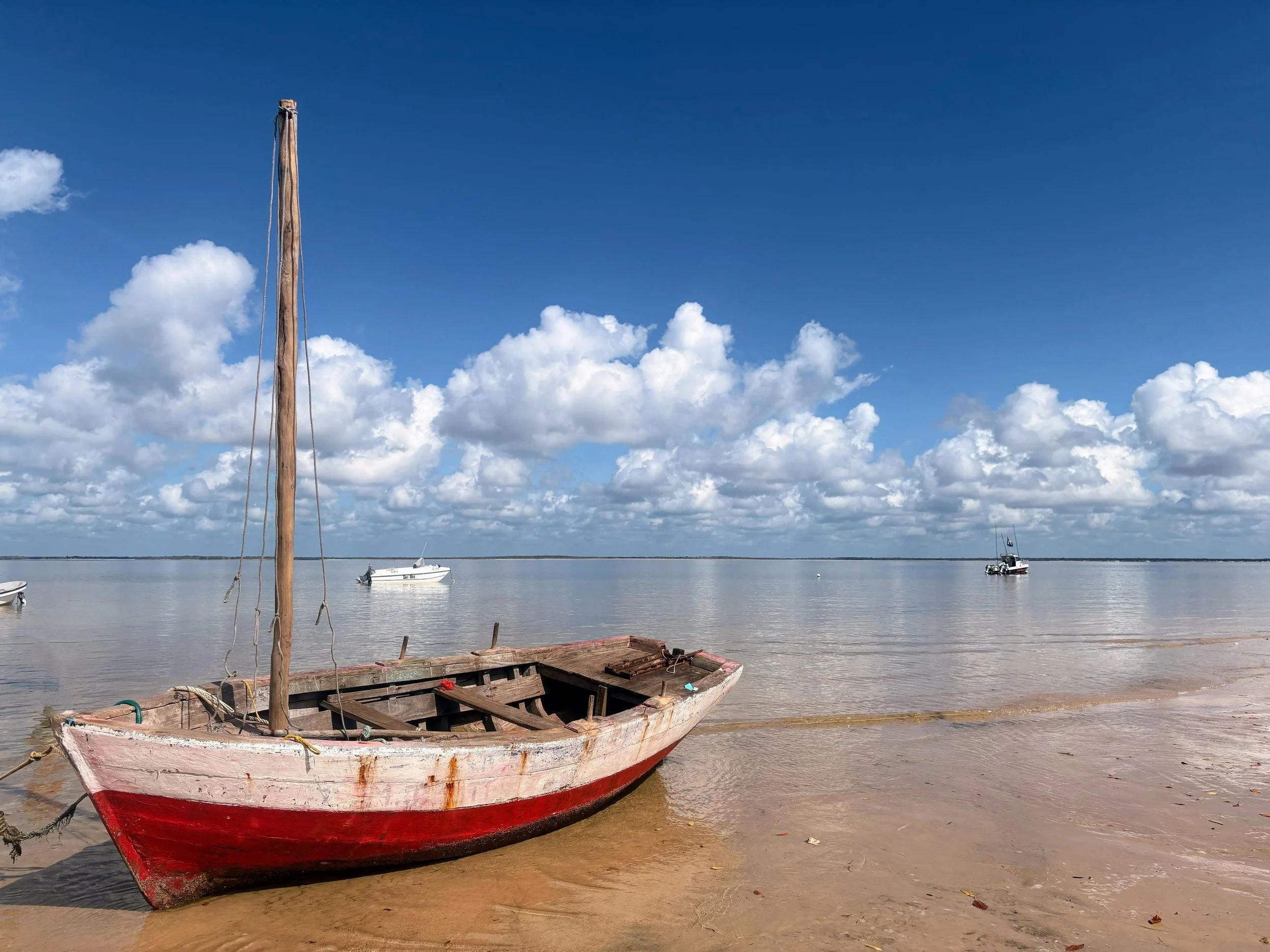 Old wooden boat with a red and white paint job resting on a sandy beach, with a tall wooden mast but no sails. The water is calm with a few boats and a cloudy sky in the background.