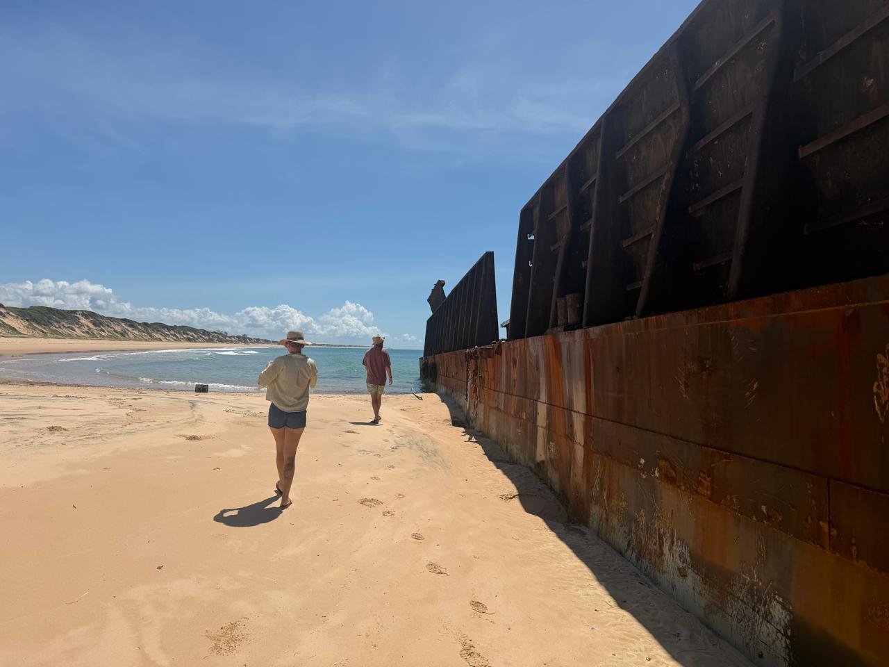 Two people walking along a sandy beach near an old, rusted shipwreck under a clear blue sky.