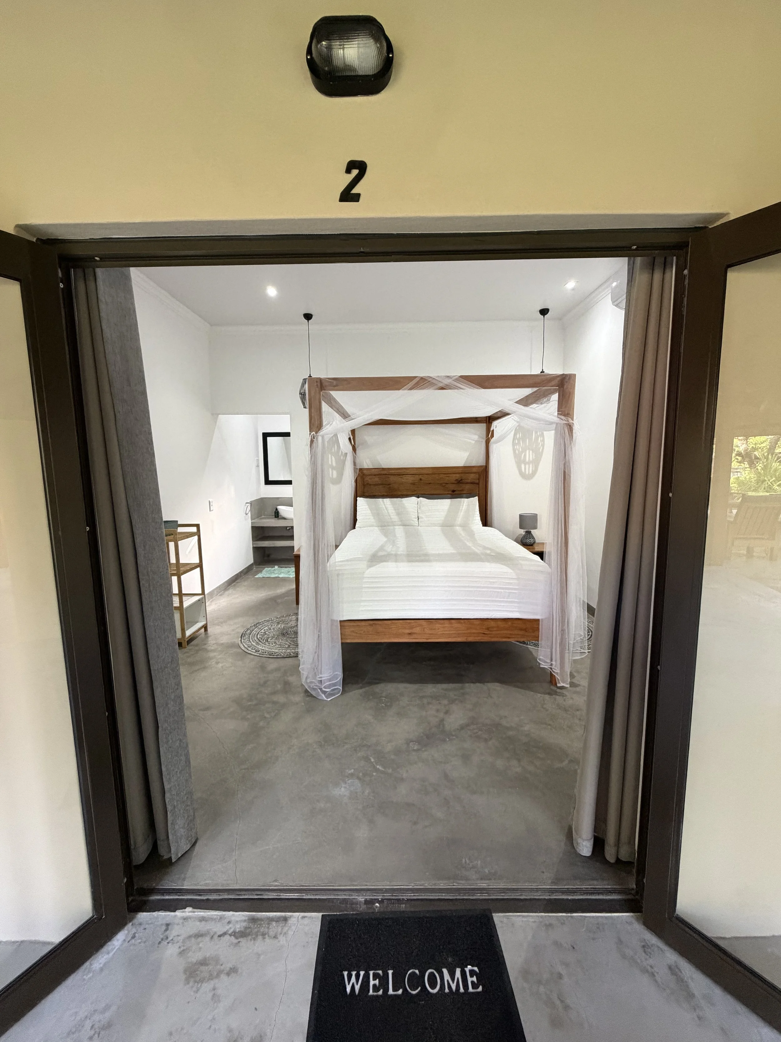 Open glass doors leading into a hotel room with a welcome mat on the floor. Inside, there is a bed with a wooden headboard and sheer curtains, side tables with lamps, and a small seating area with a wooden table.