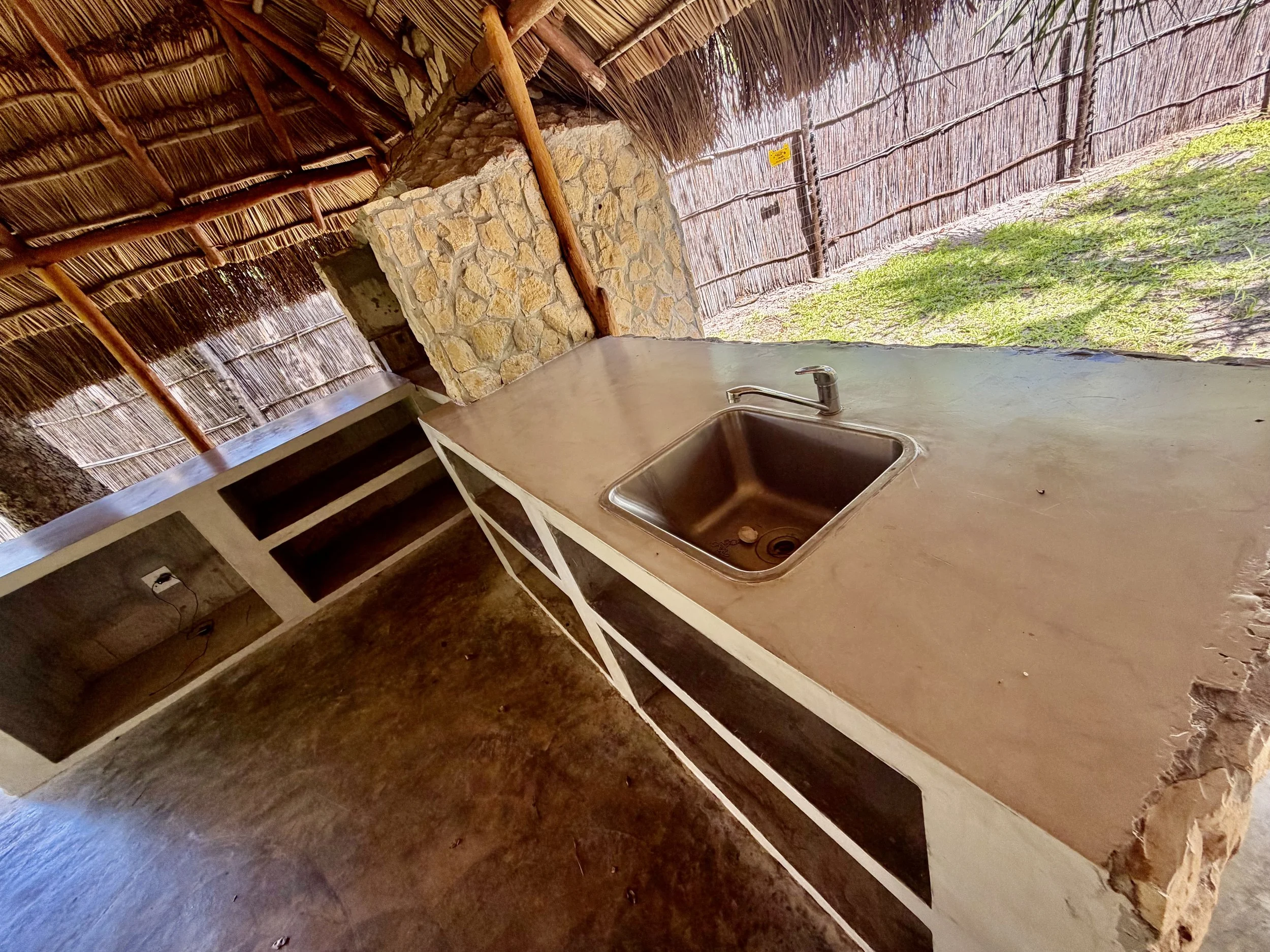 Wooden counter with a stainless steel sink, stone chimney, bamboo fencing, and a thatched roof. Bright outdoor grass is visible.