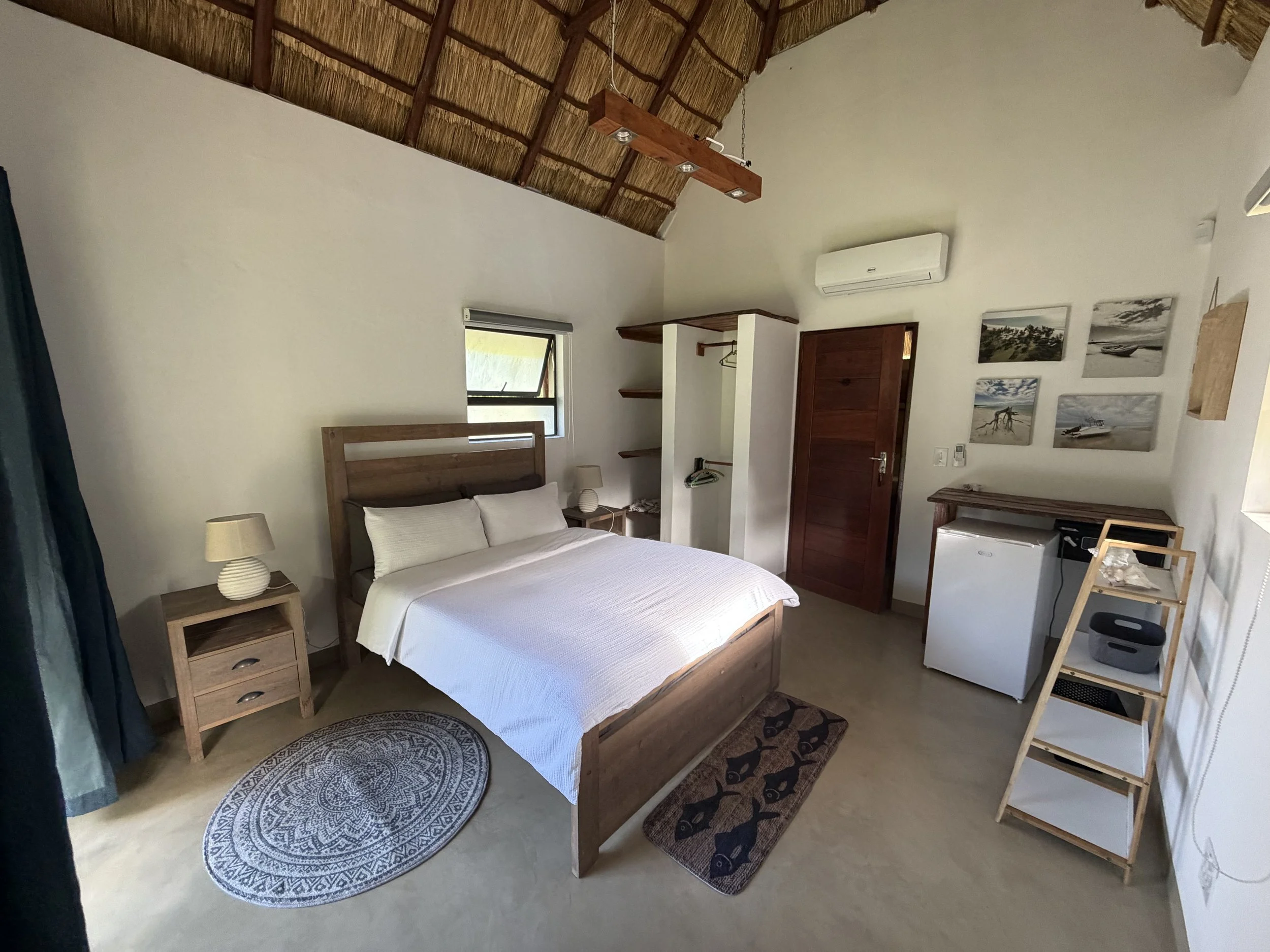 A bedroom with a wooden bed frame and white bedding, two matching bedside tables with lamps, a circular rug, a small closet with open shelves and a hanging rod, a window with a shade, and wall art featuring beach scenes, a mini-fridge, and a wooden ladder shelf.