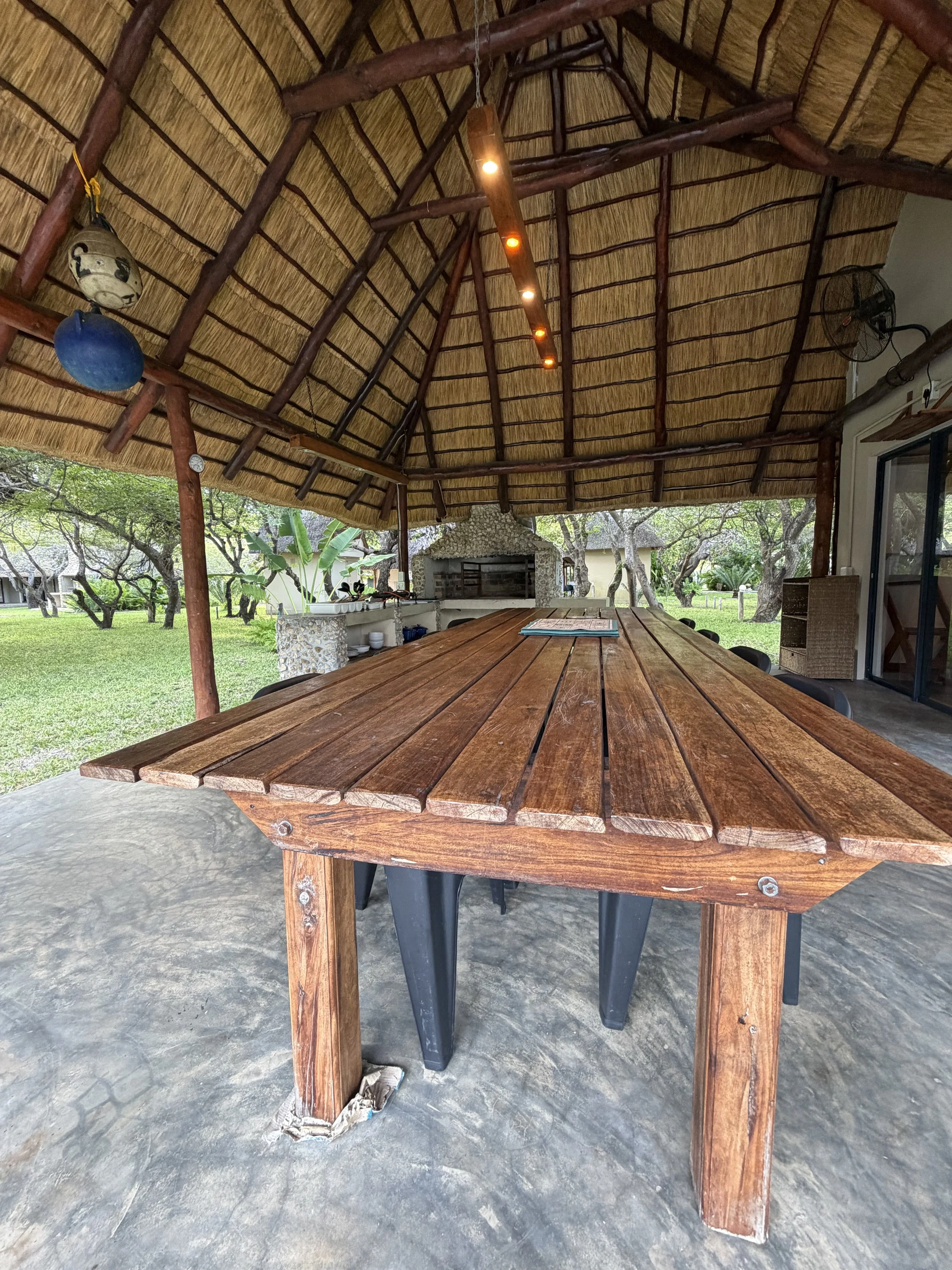 Outdoor covered dining area with a long wooden table and black plastic chairs, bamboo roof with wooden support beams, and a built-in stone barbecue grill in the background, surrounded by green trees and grass.
