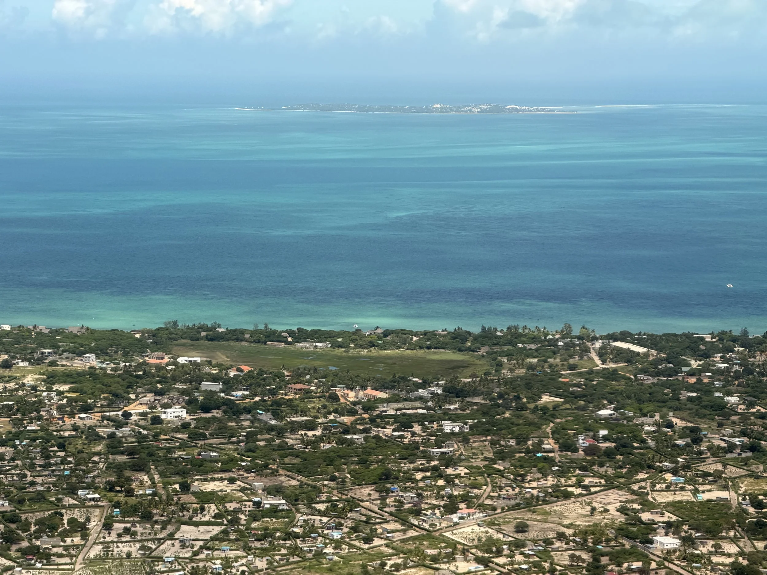 A coastal landscape with a small city or neighborhood in the foreground, lush greenery, and an expansive body of water with a distant landmass in the background under a partly cloudy sky.