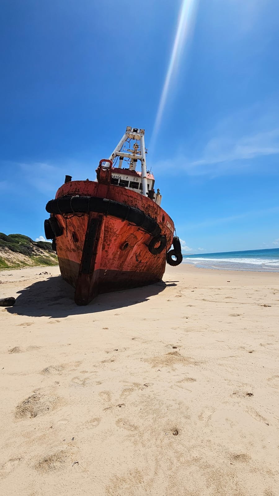 An old, rusty red boat resting on a sandy beach with footprints, facing the ocean under a bright blue sky.