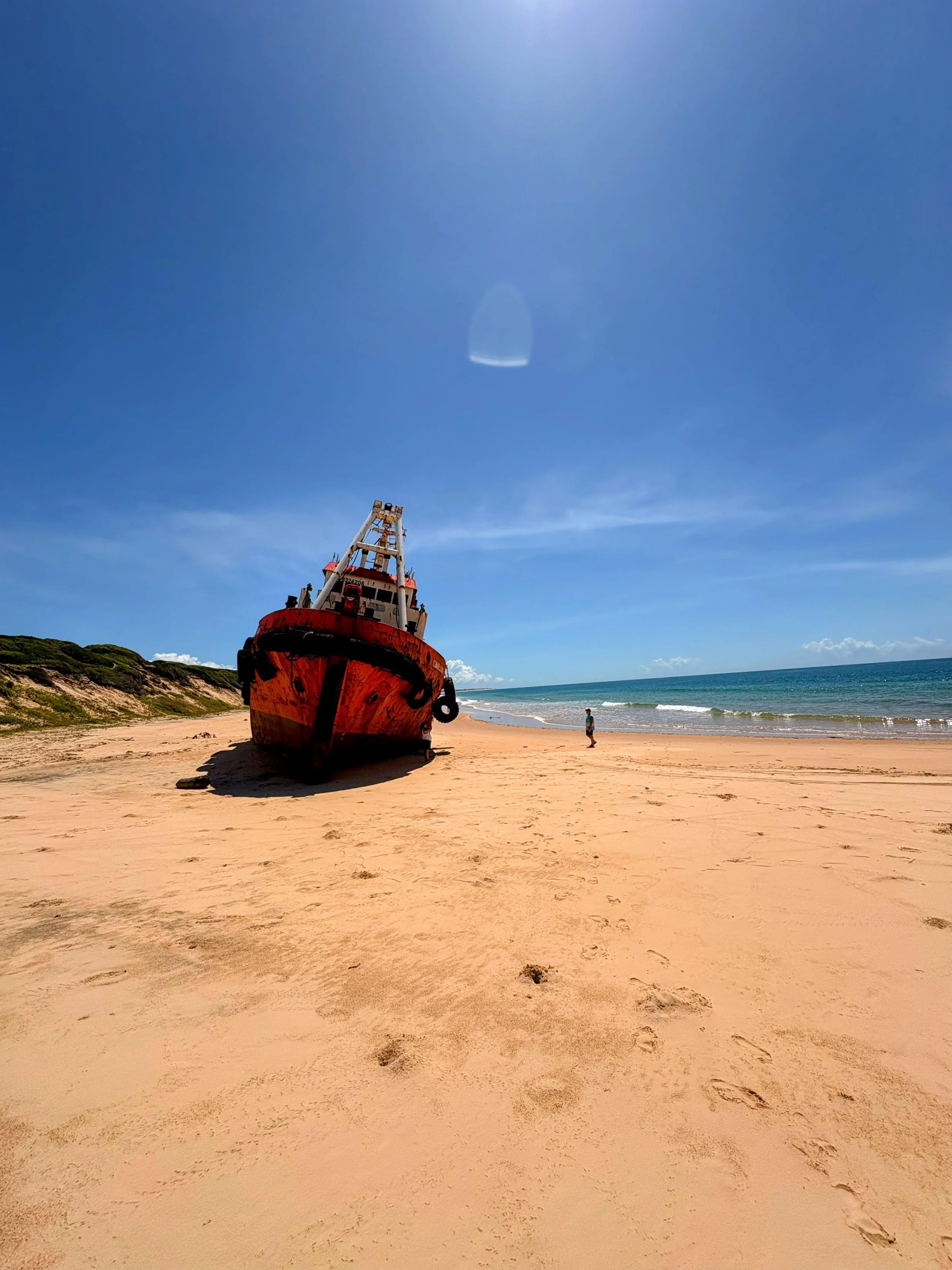 A large, rusty red shipwreck on a sandy beach with a person walking nearby, ocean waves, and a clear blue sky overhead.