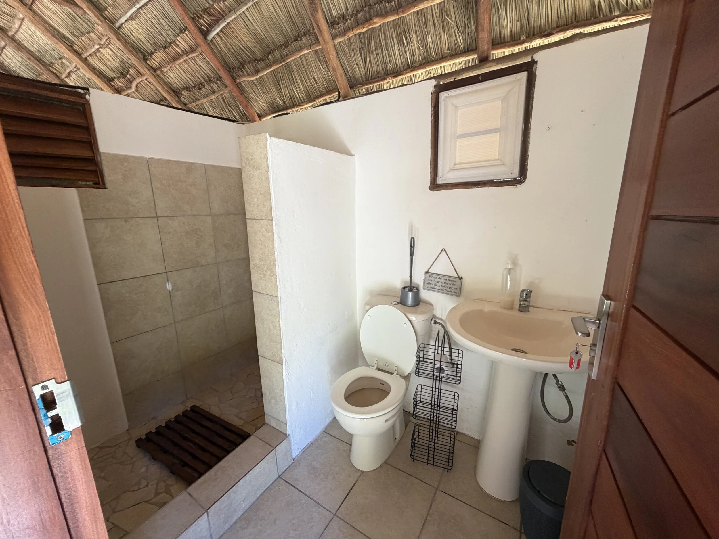 A small bathroom with a toilet, a pedestal sink, and a shower area with beige tiles and a wooden slatted shower mat. There is a small window with a wooden frame on the white wall above the toilet. The ceiling appears to be made of thatch or bamboo, giving it a rustic appearance.