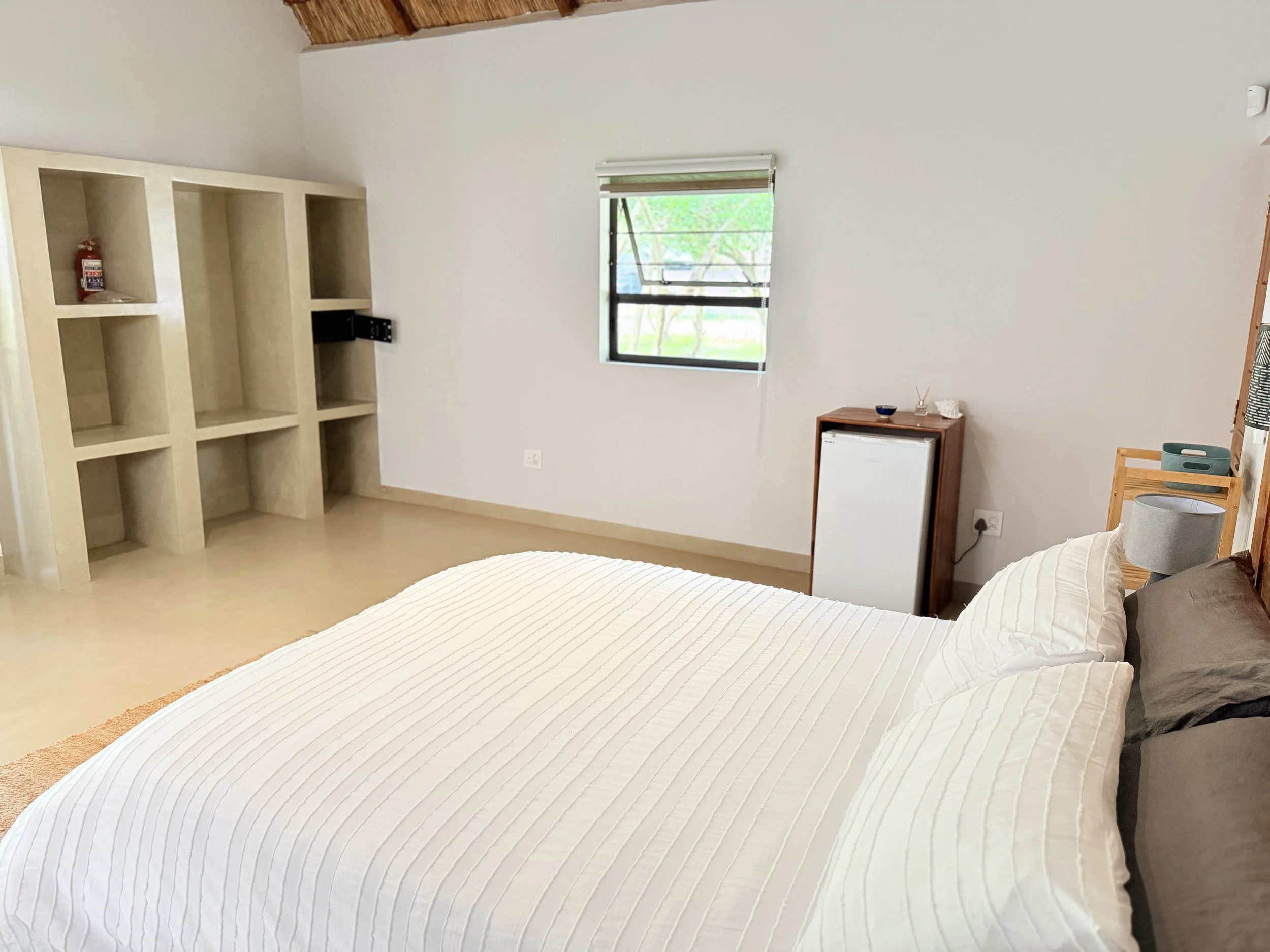 A bedroom with a bed, a wooden shelf, a small refrigerator, and a window with a view of trees outside.