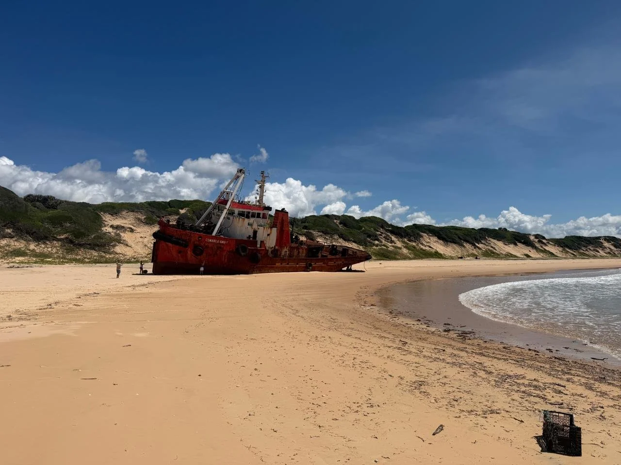 A large, rusted ship stranded on a sandy beach with green, hill-like dunes and a partly cloudy blue sky in the background.