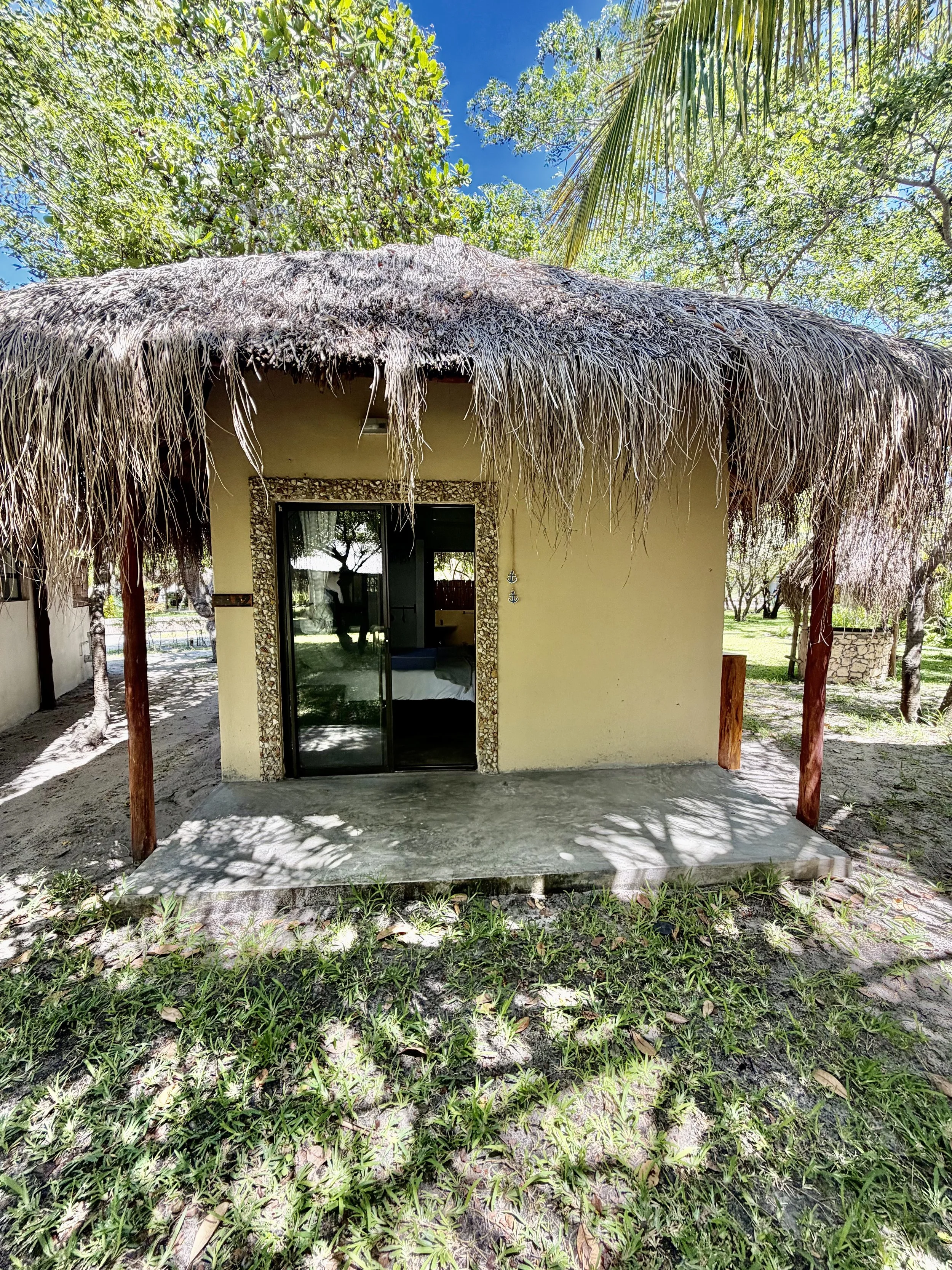 A thatched-roof hut with an open door, surrounded by tropical trees and grass, under a blue sky.