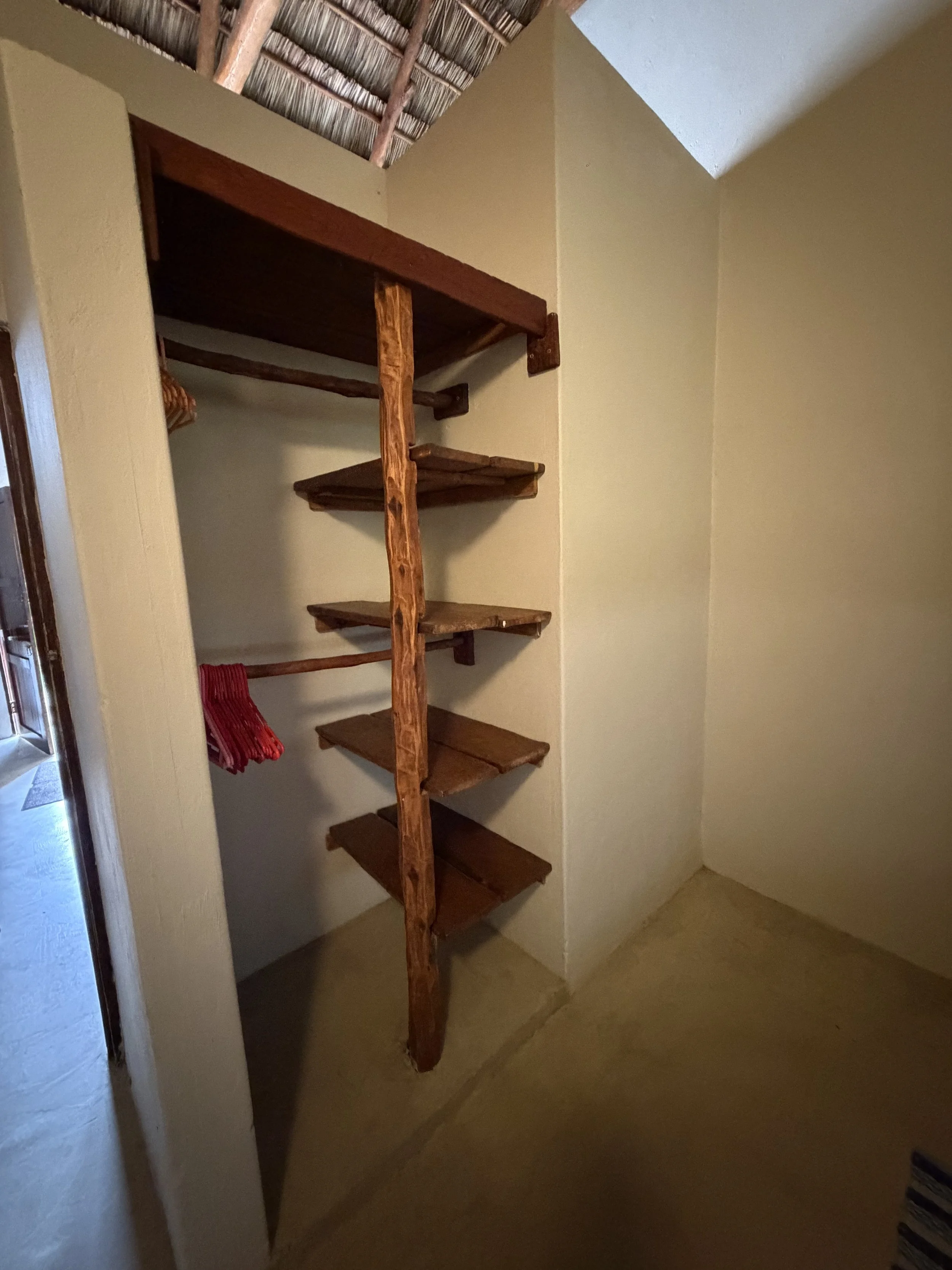 A rustic wooden staircase with open shelves on the side, leading up to a thatched roof ceiling inside a room with beige walls and a beige tiled floor.