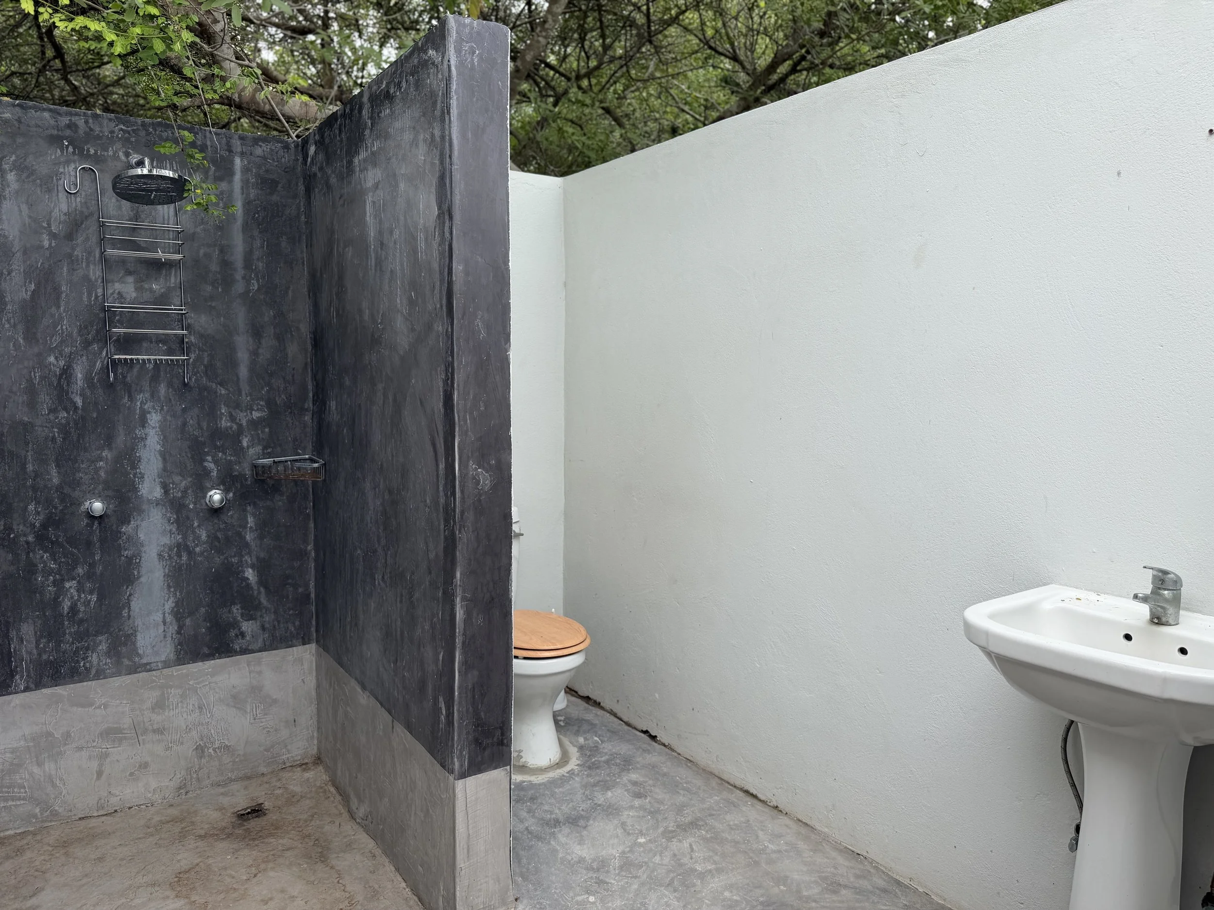 An outdoor bathroom with a black cement wall that has a showerhead and a small shelf, next to a white wall with a toilet and a white sink. There are trees visible above.