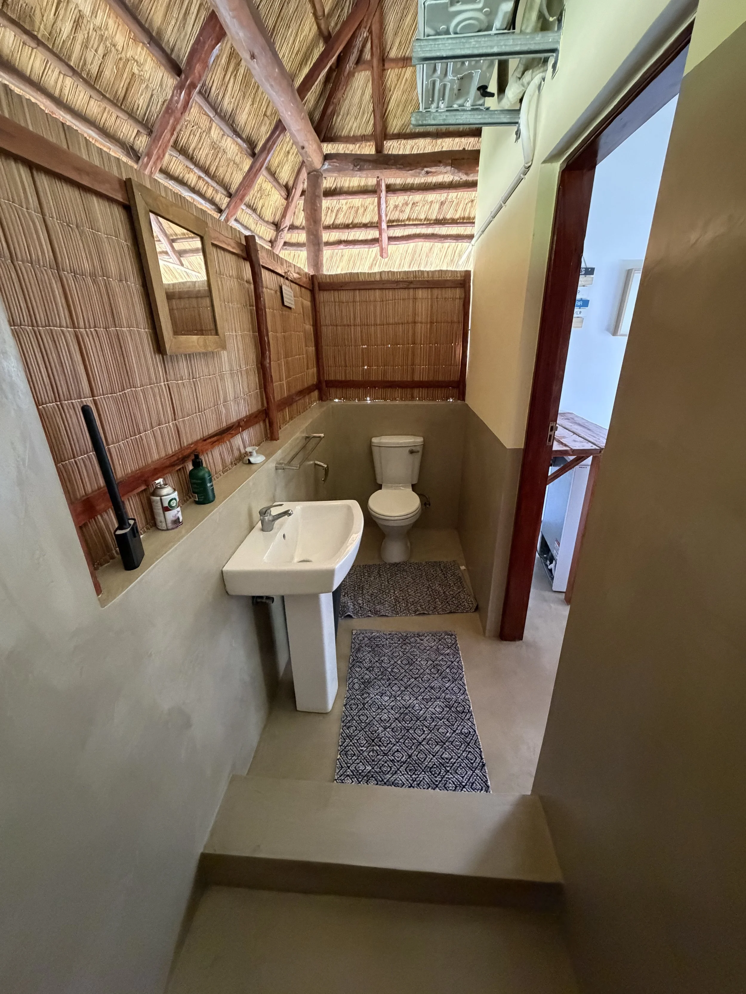 A small bathroom with a rustic style. It has a white toilet, a ceramic sink with a silver faucet, two patterned rugs on the beige floor, and a bamboo wall with a mirror. The bathroom features a thatched roof and wooden support beams, with some shelves and cleaning items placed on a ledge.