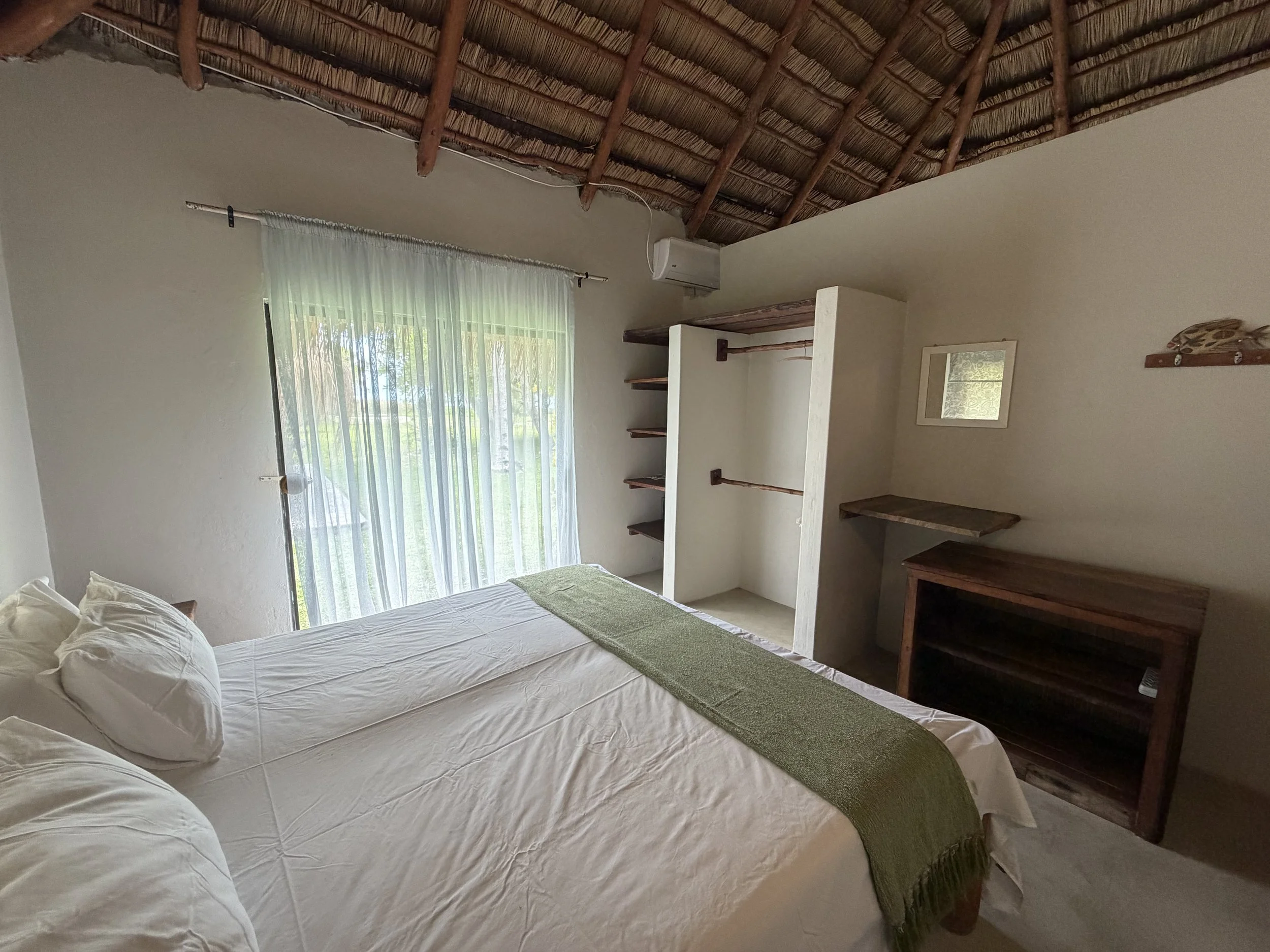 A bedroom with a bed covered in white sheets and pillows. There is a green runner on the bed. A large window with sheer curtains lets in natural light. The ceiling has a thatched roof with wooden beams. There are built-in shelves and a small desk or table in the corner. An air conditioning unit is mounted on the wall.