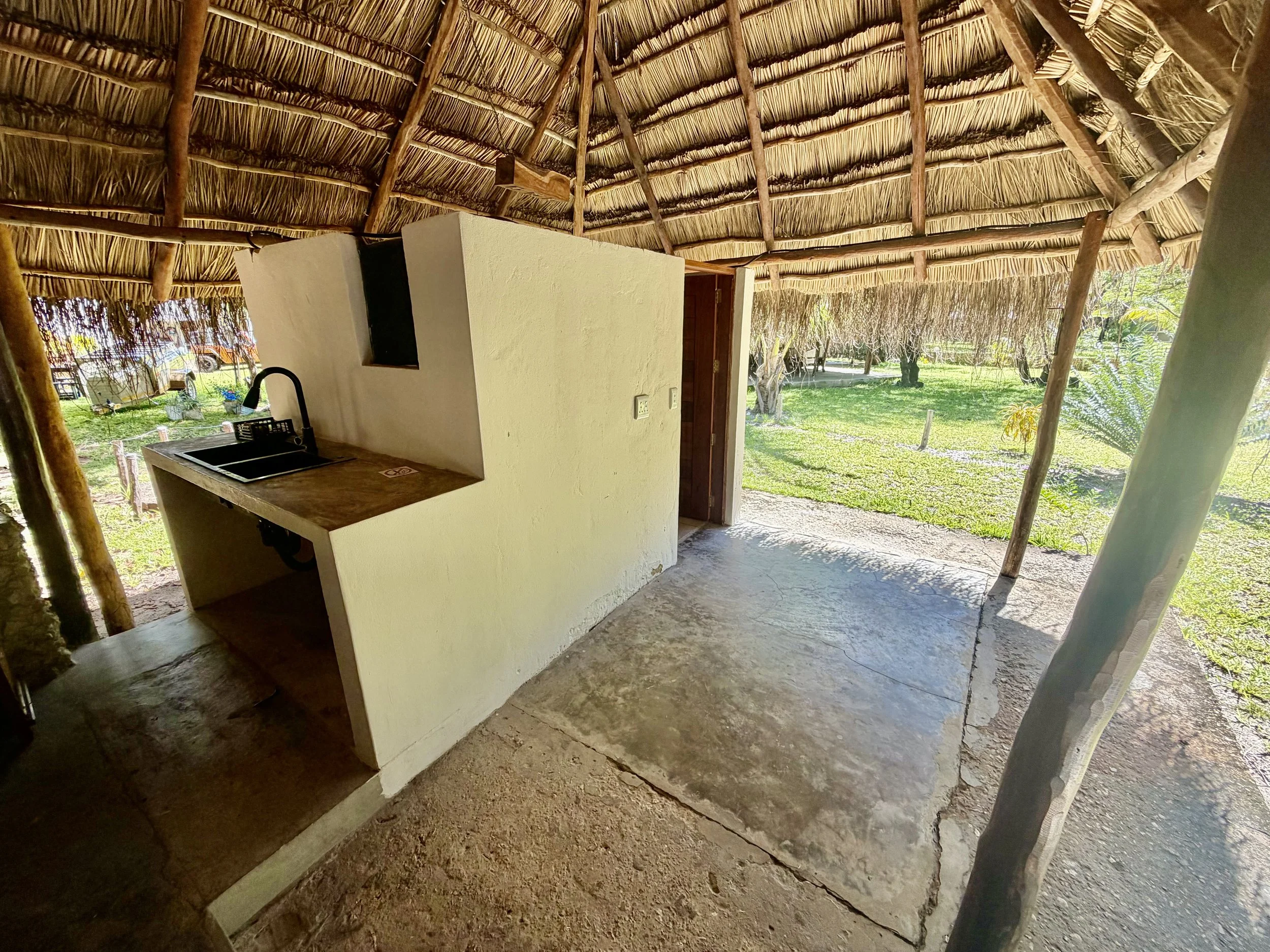 View inside a thatched hut with a small white wall, a corner sink with a black faucet, and a door opening to a grassy outdoor area with trees.