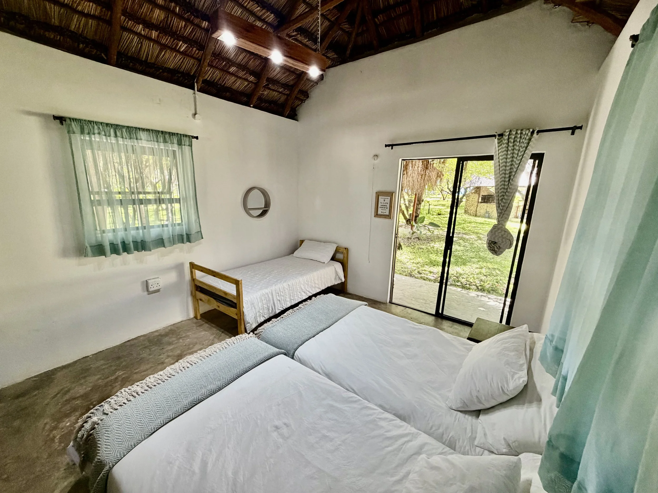 A bedroom with three neatly made white beds, a window with light green curtains, and a sliding glass door leading outside to a grassy area with trees. The ceiling is made of thatch with wooden beams.
