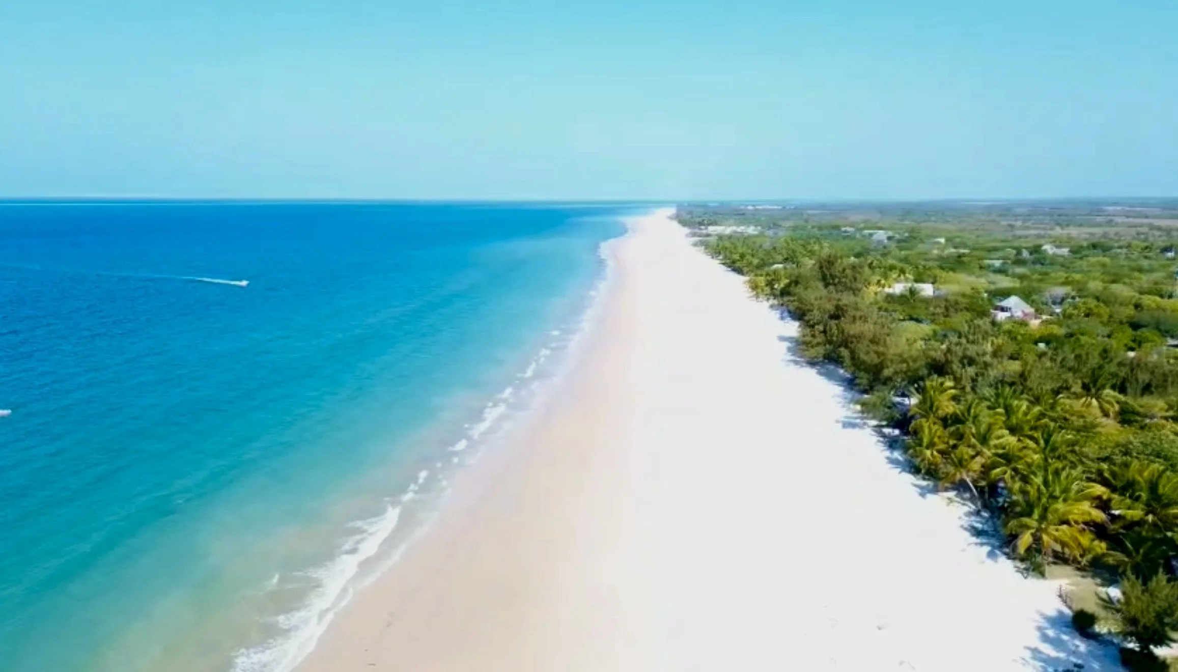 Aerial view of a tropical beach with turquoise water and white sand, lined with green trees and small houses in the background.