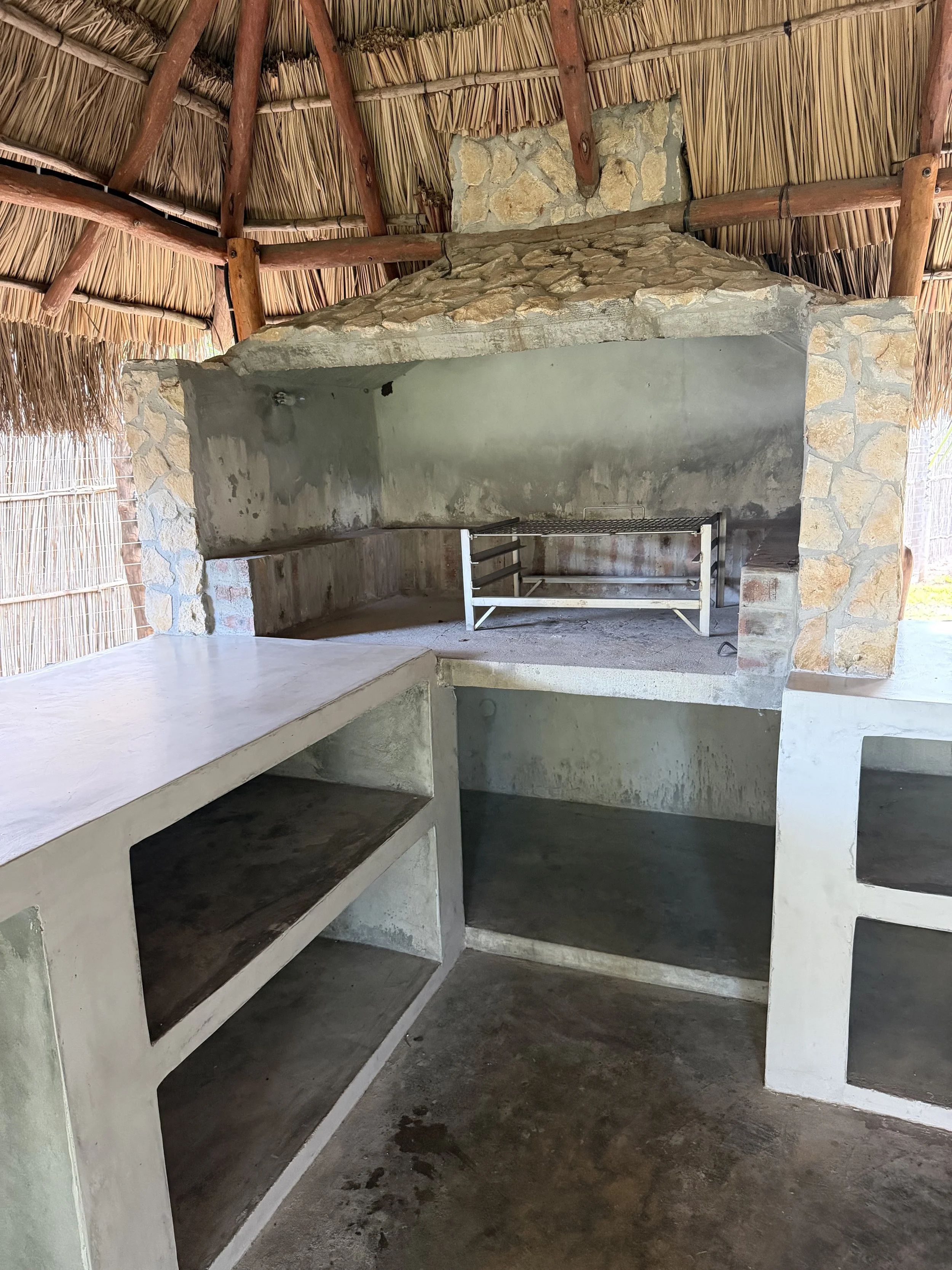 A rustic outdoor kitchen area with stone and concrete structure, a grill, and a thatched roof supported by wooden beams.