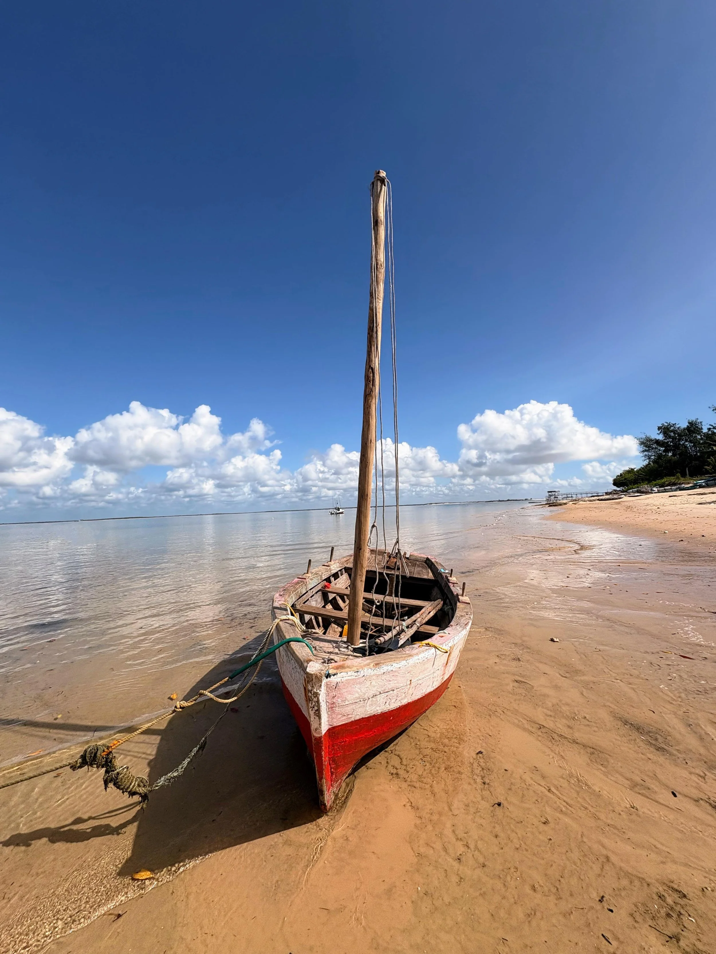 A weathered small boat with a single mast is stranded on the sandy shore, with the calm water gently lapping at its side. The sky is mostly blue with some scattered clouds, and there are trees and structures visible in the distance.